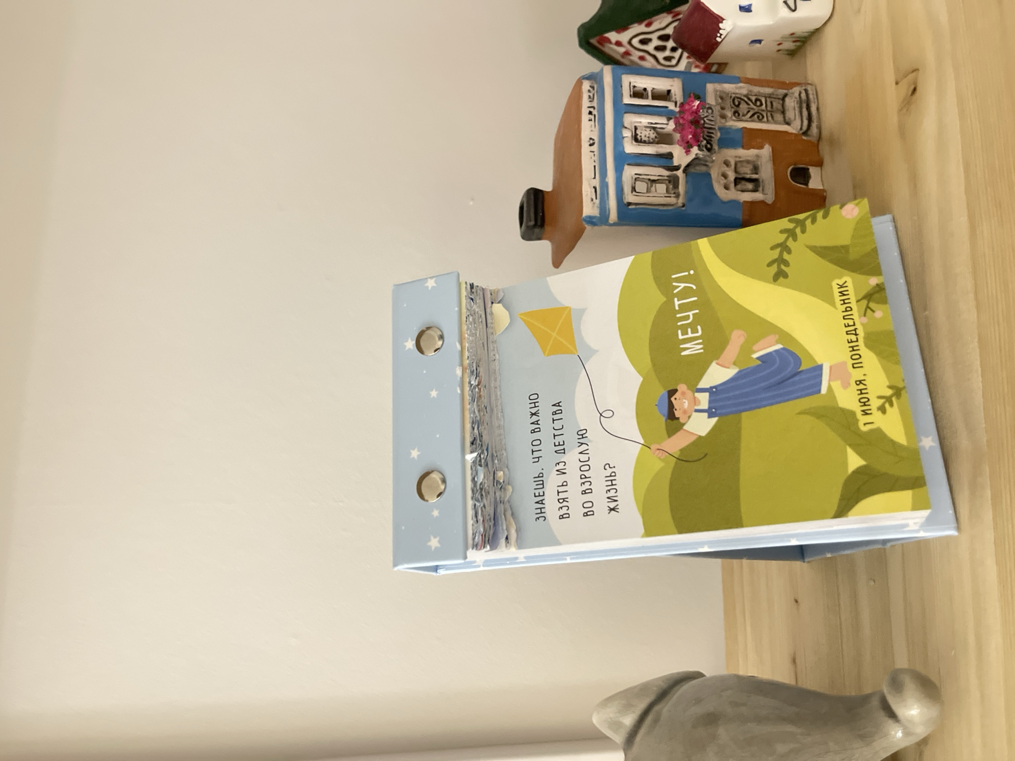 Here is a detailed description of the image:
The image shows a children's book titled "МЕЧТУ!" (Dream!) propped open on a light wooden surface, possibly a shelf. The book cover features an illustration of a child in a blue striped outfit flying a yellow kite against a backdrop of green hills. To the left of the illustration is text in Russian that reads: "ЗНАЕШЬ, ЧТО ВАЖНО ВЗЯТЬ ИЗ ДЕТСТВА ВО ВЗРОСЛУЮ ЖИЗНЬ?" (You know what's important to take from childhood into adulthood?). The lower right corner of the book cover indicates "1 ИЮНЯ, ПОНЕДЕЛЬНИК" (June 1st, Monday). The book has a light blue cover with white stars and three metal rings along the spine.
Behind the book, there are decorative miniature houses, one primarily blue and another with a green roof. Below the book is a grey object with a knob-like shape. The background consists of a plain white wall. The photo is slightly tilted, providing a unique perspective of the objects.