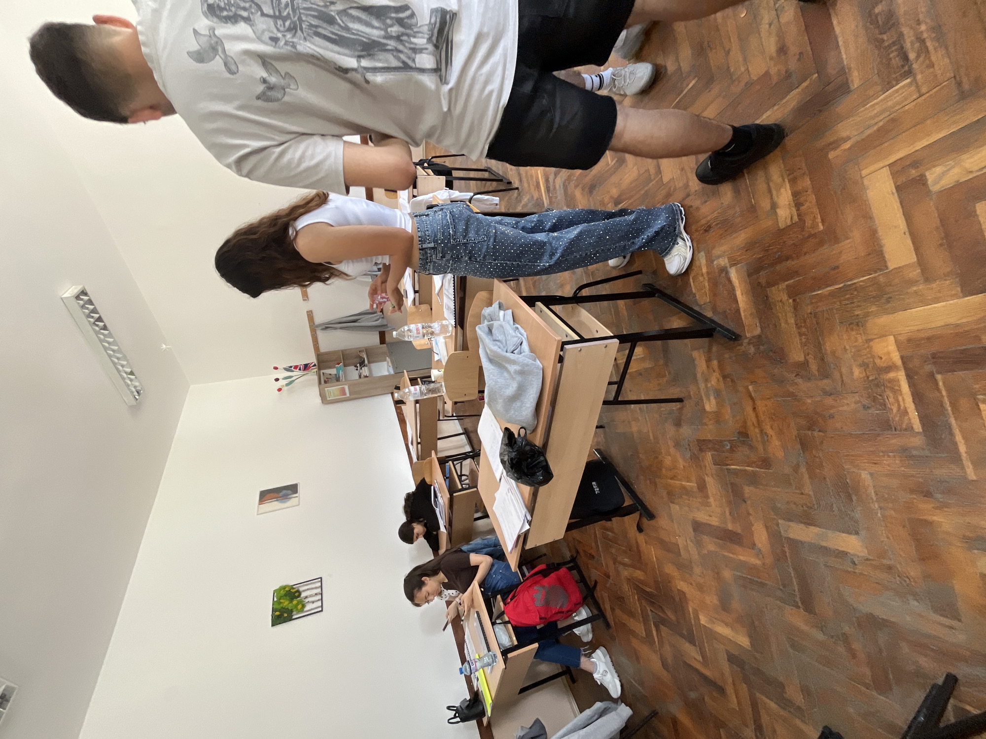 The image depicts a classroom scene, likely in Zlatograd, Bulgaria, during what appears to be daytime. The room features a patterned wooden parquet floor and plain white walls. Several individuals, presumably students, are present.
In the foreground, a male student stands with his back to the camera, wearing a white t-shirt with a gray graphic design of birds, black shorts, and black sneakers. Adjacent to him, a female student stands beside a wooden desk, also facing away from the camera. She wears a white tank top and blue jeans adorned with numerous white embellishments or sequins, paired with white sneakers.
Further into the room, two female students are seated at individual wooden desks with black metal frames. They are bent over their desks, seemingly engaged in writing or studying. One seated student wears a dark top and blue jeans, with a red bag placed beside her desk. Underneath her desk, a black bag has the text "EZEL" visible. A clear water bottle, possibly with a blue cap, is on one of the desks. Papers, bags, and a gray sweatshirt are also visible on or near the desks.
The classroom is brightly lit, with a rectangular fluorescent light fixture mounted on the white ceiling. The walls are sparsely decorated with two small framed items: one appears to be a landscape painting, and the other a green, plant-like decoration within a black frame. A wooden shelf unit with various small objects and a miniature flag-like decoration is seen on the wall in the background. The overall atmosphere suggests a typical school environment.