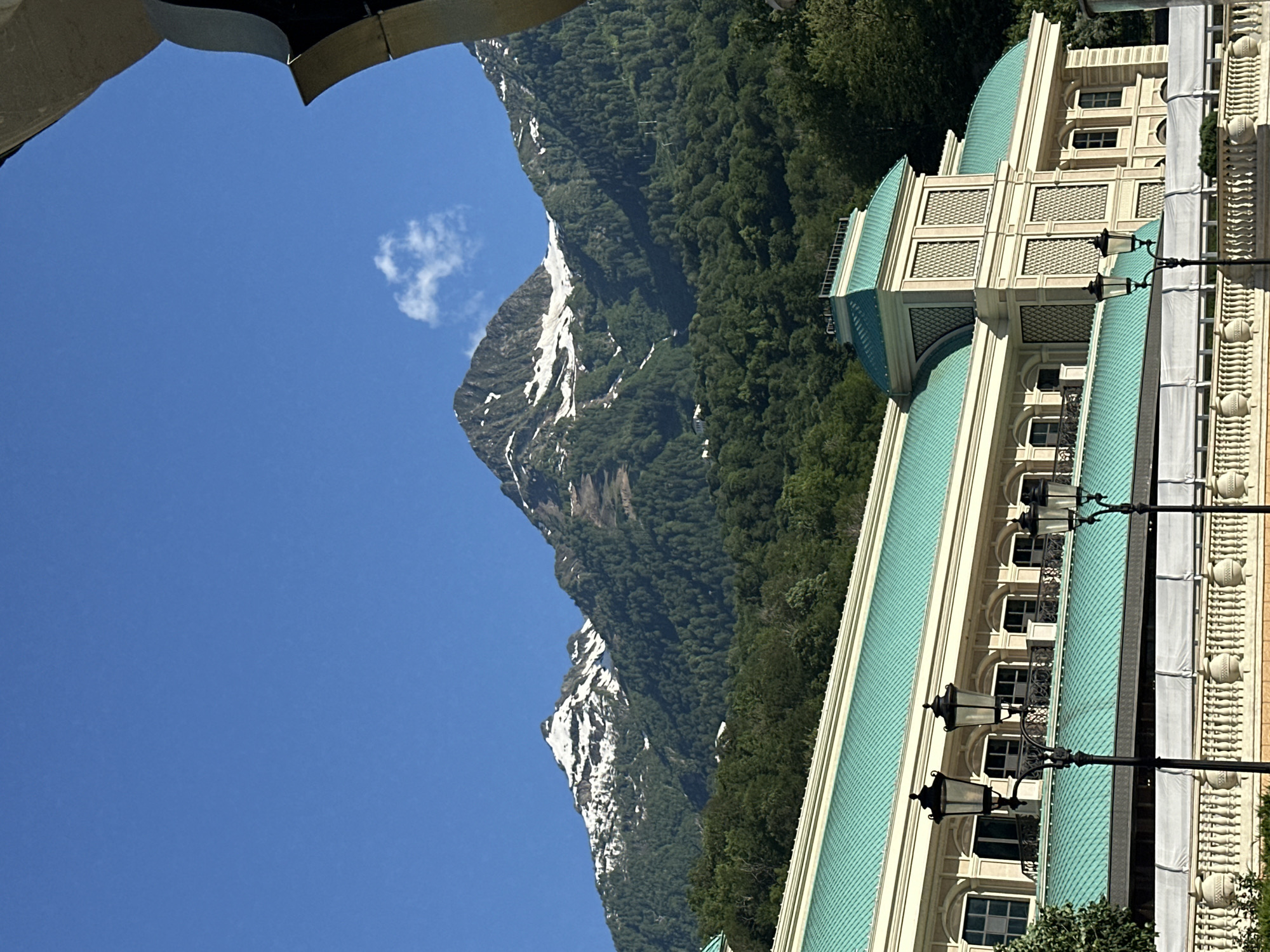 The image captures a scenic view of the mountains behind a building with a turquoise roof in Estosadok, Russia. In the foreground, a light-colored building with classical architectural elements and a distinctive turquoise roof dominates the lower half of the frame. Ornate lampposts stand in front of the building. Behind the building, lush green forests cover the lower slopes of imposing mountains. The peaks of these mountains are capped with snow, suggesting a high altitude. The sky above is a clear, vibrant blue with a few wispy clouds. The composition suggests a bright, sunny day.