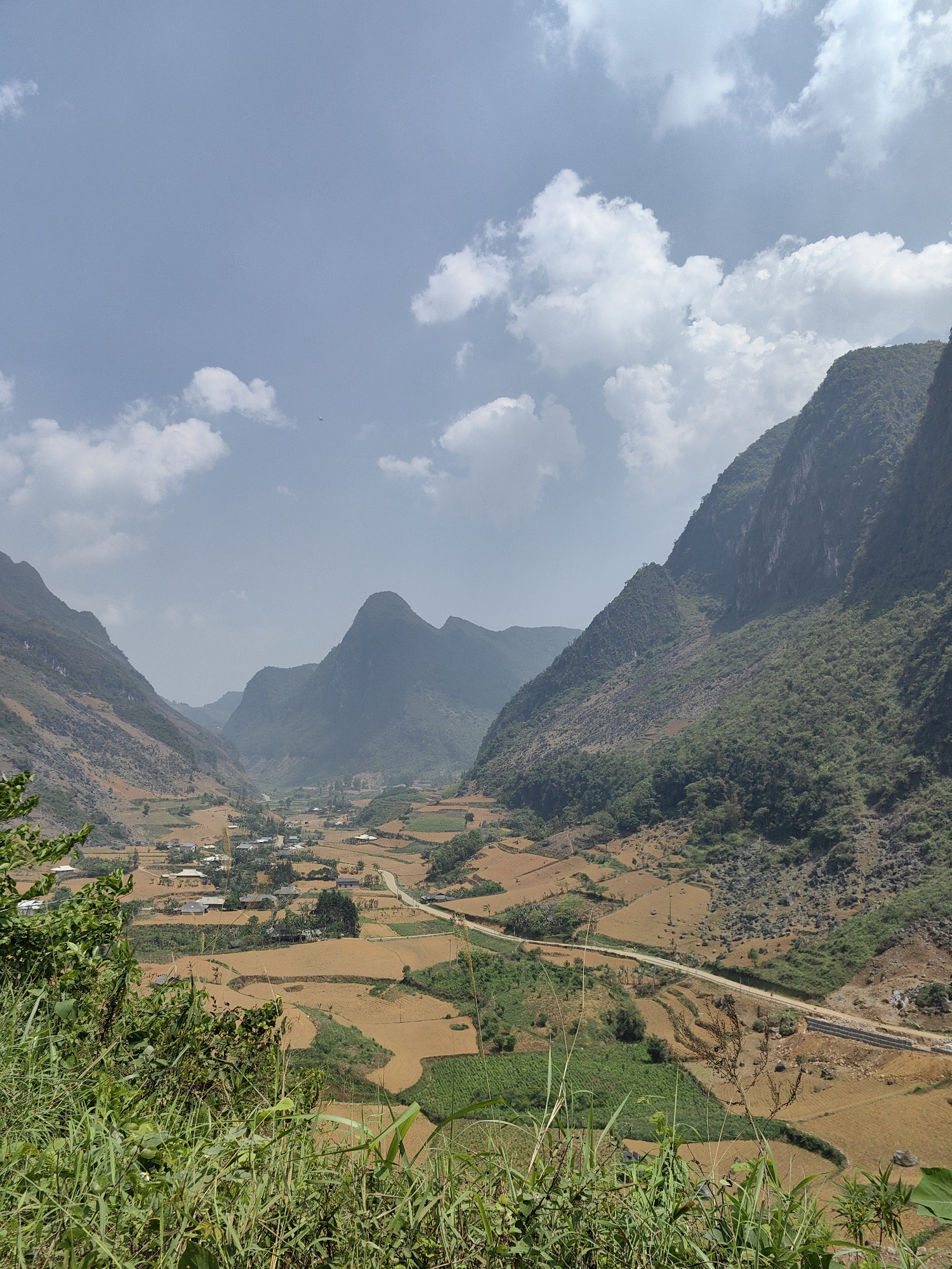 The image depicts a wide, deep valley in Xã Xuân Trường, Vietnam, dominated by towering mountains on both sides. The valley floor is a patchwork of brown and green fields, interspersed with small clusters of buildings that form a village. A road cuts through the valley floor. The sky is partly cloudy. Foreground vegetation is visible.