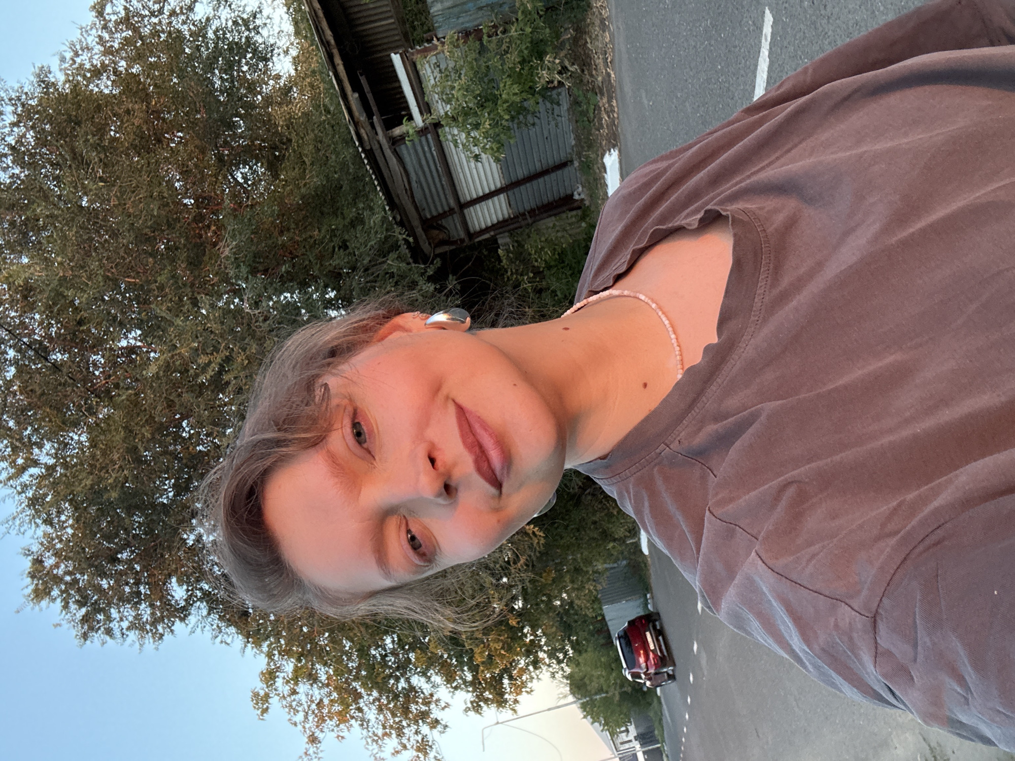 A selfie taken outdoors shows a woman from the chest up. She is wearing a muted brown t-shirt and a light pink beaded necklace. She has light skin, light blue eyes, and light brown hair pulled back. She is wearing large silver hoop earrings. Her face is turned slightly to her left and she is smiling. The background includes a large tree with green leaves, a rustic corrugated metal structure to her right, and a paved road with a white line on the left. A red car is visible on the road in the distance. The sky is a clear blue. The lighting suggests it is either sunrise or sunset, with warm light illuminating the woman's face and the surrounding environment. The location is Aksay, Kazakhstan.
