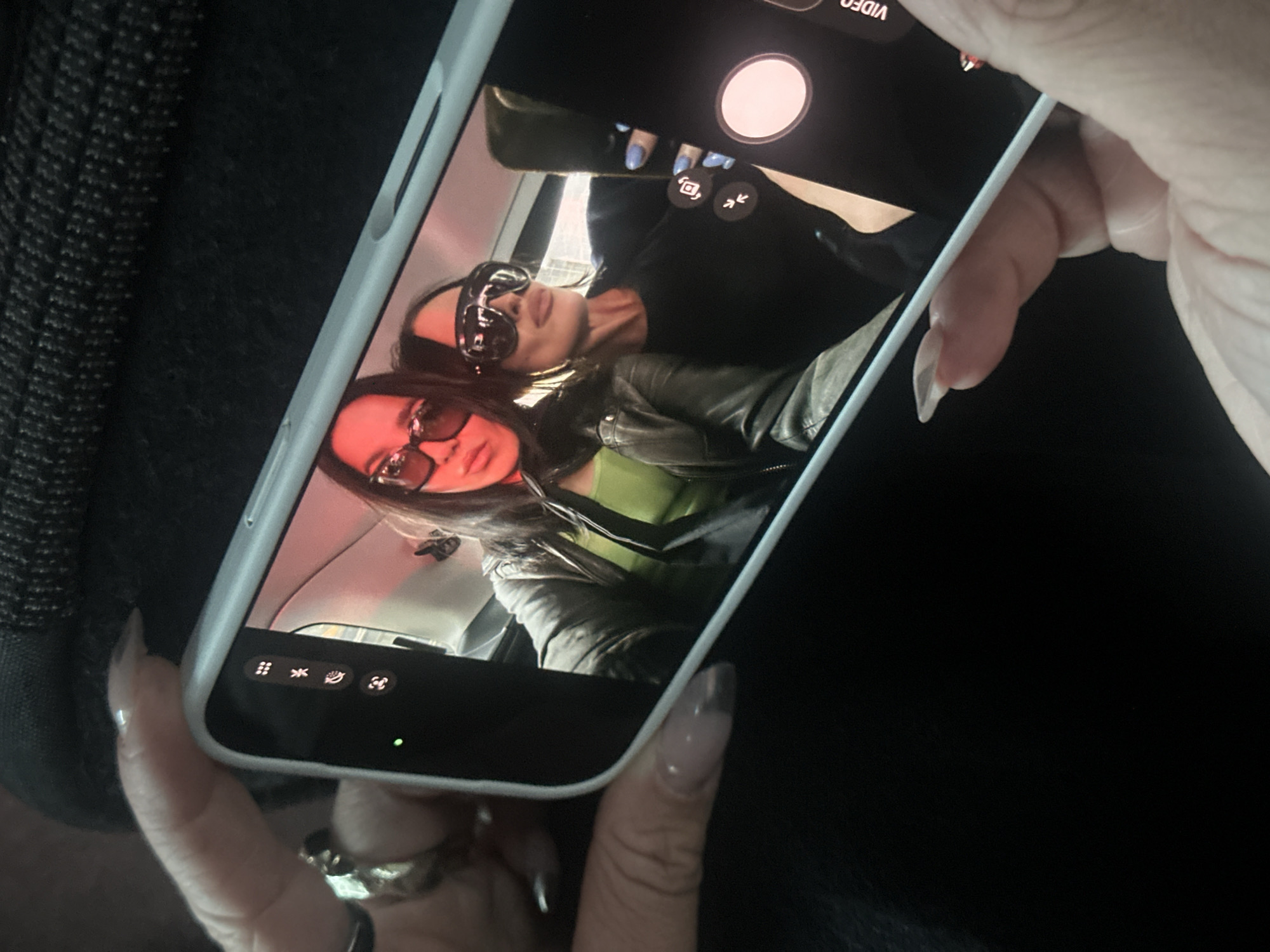 A person's hands are shown holding a white smartphone, likely an iPhone, in an angled shot against a dark, possibly textured fabric background. The person holding the phone wears at least one prominent ring on a finger, and their nails are long and clear-polished.
The smartphone screen displays a photograph or live view from the camera application, featuring two women. Both women have dark hair and are wearing sunglasses, obscuring their eyes. The woman closer to the top of the screen is wearing a black top and has bright blue nail polish on her fingers. The second woman, slightly lower on the screen, is wearing a vibrant green top under a dark leather jacket. The setting for the two women within the phone screen appears to be an indoor space or a vehicle interior, possibly a car, indicated by light-colored upholstery behind them.
The phone's camera interface is visible on the screen, including a prominent white circular shutter button, the partially visible word "VIDEO" at the top, and various smaller icons for camera settings such as flash, grid, live photo, timer, and camera flip/switch. The image captures a casual moment of someone reviewing or taking a picture of two stylish individuals. The location context for this scene is London, United Kingdom.