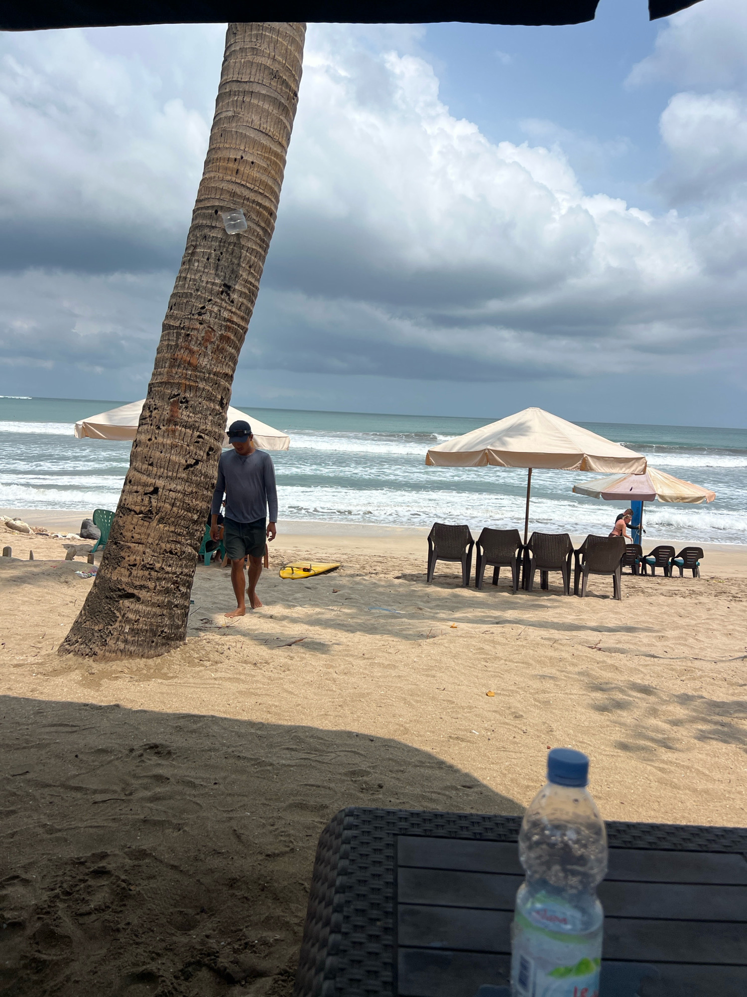 The image depicts a beach scene in Kuta, Indonesia. In the foreground, a bottle of water sits on a dark, woven surface. The beach is sandy, with a prominent palm tree on the left side. A man wearing a grey long-sleeved shirt, dark shorts, and a blue cap is walking on the sand near the palm tree. In the middle ground, beach chairs and umbrellas are set up. The ocean with waves is visible in the background, under a cloudy sky. A yellow surfboard is lying on the sand near some green plastic chairs.