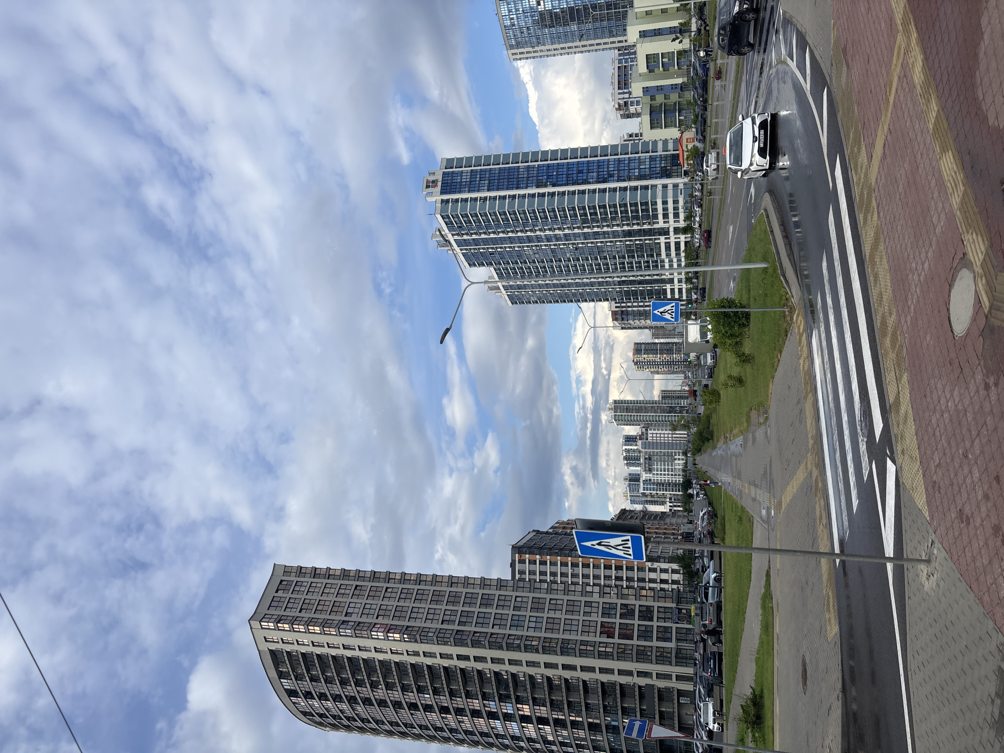 The image captures a street scene in the Senitsky Sel'skiy Sovet, Belarus, dominated by modern, high-rise apartment buildings. On the left, a distinctive curved, grey building with numerous windows stands prominently. To the right, two taller, slender buildings with blue and white facades are visible, with more buildings stretching into the distance. The sky is overcast with a mix of white and grey clouds. A wet asphalt road curves through the scene, with a white car turning. A pedestrian crossing with white stripes is in the foreground, with red and grey paving stones. Traffic signs, including a blue sign with a white pedestrian symbol, are present. Cars are parked along the roadside, and a few pedestrians are seen walking on a sidewalk. The overall impression is an urban environment with contemporary architecture under a cloudy sky.