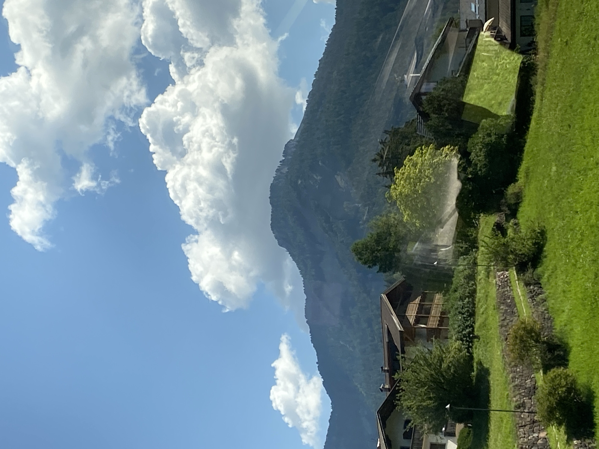 The image depicts a scenic mountain landscape with a village nestled at its base. In the foreground, a vibrantly green, grassy hill slopes upwards, dotted with small bushes. A terraced stone wall runs horizontally across the middle ground, further enhancing the stepped terrain. Several traditional alpine houses with wooden balconies and dark roofs are visible, surrounded by lush trees and dense foliage. Tall lampposts stand intermittently along the pathways.
Dominating the background is a majestic, tree-covered mountain peak, its slopes partially obscured by shadows. The sky above is a clear, bright blue, adorned with fluffy white cumulus clouds. The overall impression is one of serene natural beauty and traditional alpine living, likely in a location like Urtijëi - St. Ulrich in Gröden - Ortisei, Italy, a region known for its picturesque mountain villages and stunning natural scenery. There are no people or animals visible in the image.