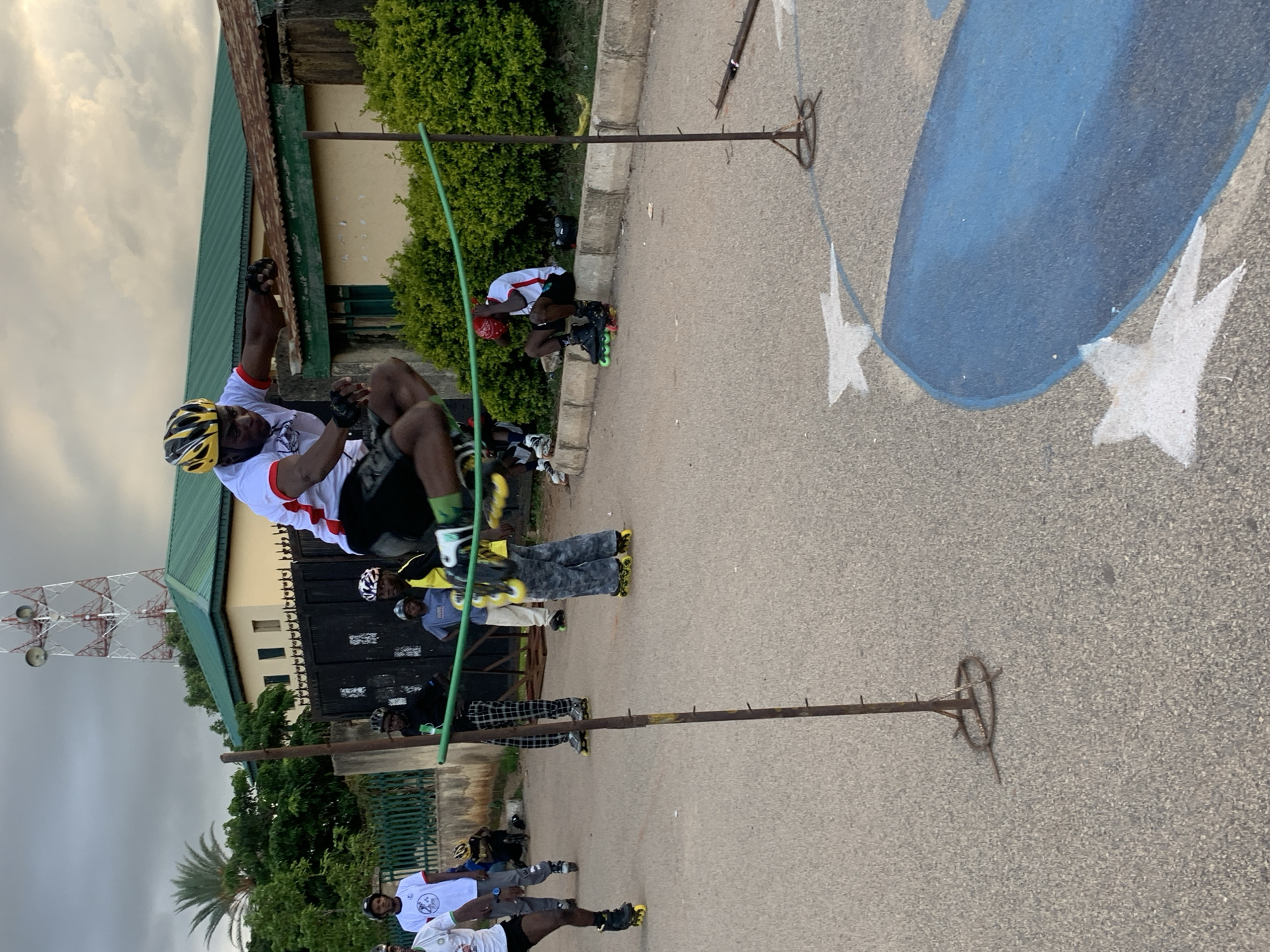 In Jos, Nigeria, a group of individuals are participating in an inline skating event. The central focus is a young man in mid-air, actively jumping over a green cord stretched between two poles. He is wearing a yellow and black helmet, a white t-shirt with red trim, black shorts, and inline skates. Several other individuals are present, some on skates, others observing. One person is seated on a curb, and another is standing near a fence. The background features a building with a green roof, a large communication tower, and lush green trees. The ground is paved, with white chalk markings of stars and an abstract blue shape visible. The sky is overcast with clouds.
