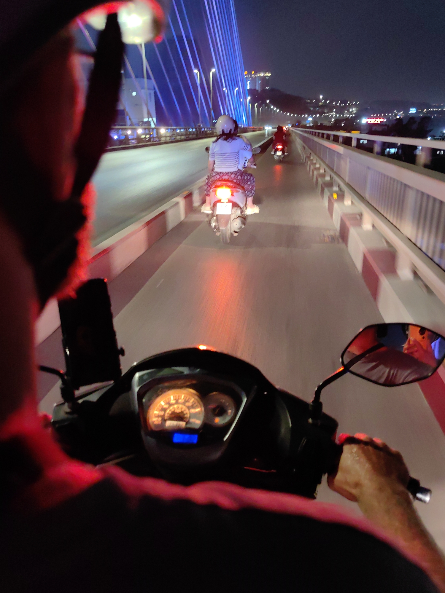 A nighttime, first-person view from the perspective of a motorcycle rider on a bridge in Hong Gai Ward, Vietnam. Two other motorcycles are ahead. The rider is holding the handlebars, and the dashboard is visible. The bridge has white and red barriers, and blue lights illuminate its structure. The city lights are visible in the distance, across the water. The head of the rider is illuminated by a red light source in the immediate foreground.