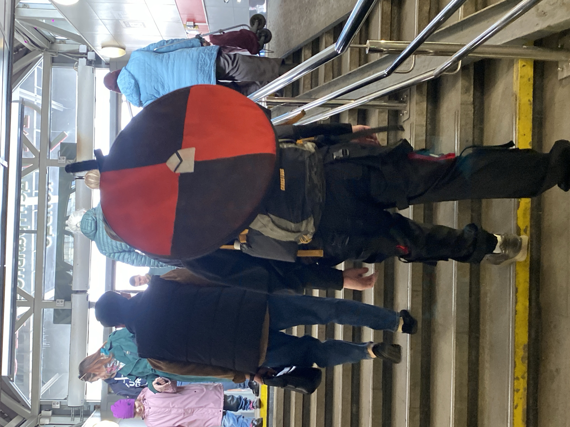 The image depicts a group of individuals ascending a concrete staircase within a pedestrian access point or transfer area of the Moscow Metro system in Moscow, Russia.
In the immediate foreground, a person is seen from behind, walking upwards. They wear black trousers, a black long-sleeved shirt, and light-colored sneakers with white socks. A grey and black backpack is worn, to which a large, circular, bi-colored object is attached, divided into a red half and a dark brown/black half, with a small, light-colored, shield-shaped emblem at its center. A black-hilted object, resembling a sword or knife, protrudes vertically from behind the person's head, secured to the backpack.
Ahead and to the left of this person, another individual ascends, wearing blue jeans, a light brown long-sleeved shirt, and a dark sleeveless vest. They hold a small black bag in their left hand and wear dark, backless shoes. Further up the stairs, a woman in a dark teal trench coat holds the hand of a child in a pink raincoat and a purple knitted hat. The child wears blue jeans and multicolored sneakers.
To the right of the central subjects, an individual in a light blue puffer jacket and dark trousers is also ascending, pulling a red wheeled trolley bag. In the background, partially obscured, another person is visible.
The staircase features grey concrete steps with metal nosing, and the bottom visible step has a yellow safety strip. A multi-barred metallic handrail runs along the right side of the stairs. Overhead, a structure of glass panels supported by metal beams allows natural light into the area, with some Cyrillic characters visible in reflection through the glass. The walls beside the stairs are light-colored. All individuals are in the process of walking up the stairs.