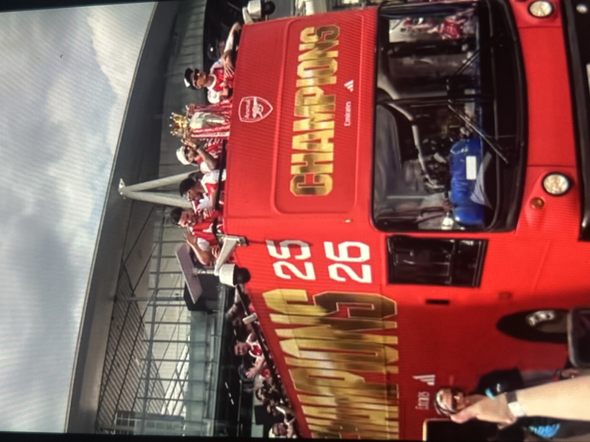 The image depicts a celebratory parade featuring a red double-decker bus carrying people, likely a sports team and staff, and a prominent trophy. The overall scene suggests a victory celebration, possibly for a football club.
On the upper deck of the bus, several individuals are visible, mostly wearing red and white jerseys, characteristic of Arsenal Football Club. One person is prominently holding aloft a large gold-colored trophy, which appears to be a cup, adorned with a smaller gold crown. Other individuals on the bus are also celebrating, some with their arms raised. The bus itself is brightly colored red and features large gold lettering.
In the background, a large, modern architectural structure with a grey facade, glass panels, and a distinctive metal framework can be seen, resembling part of a stadium or a large complex. The sky is overcast with visible clouds, indicating daytime.
Notable details on the bus include the large, bold gold word "CHAMPIONS" displayed prominently on the side and top. The Arsenal club crest, a cannon within a shield, is also visible. Below the "CHAMPIONS" text, the "adidas" logo and "Emirates" (a sponsor) are discernible. Further along the side of the bus, two large gold numbers, "25" and "26", are clearly visible, arranged vertically. Inside the driver's cab area, a blue cylinder, possibly a fire extinguisher or gas tank, can be seen. In the bottom right corner, a hand holding a smartphone suggests someone is capturing the event.
The image is rotated 90 degrees clockwise from its natural upright orientation. The general atmosphere appears joyful and celebratory, consistent with a team's championship parade.