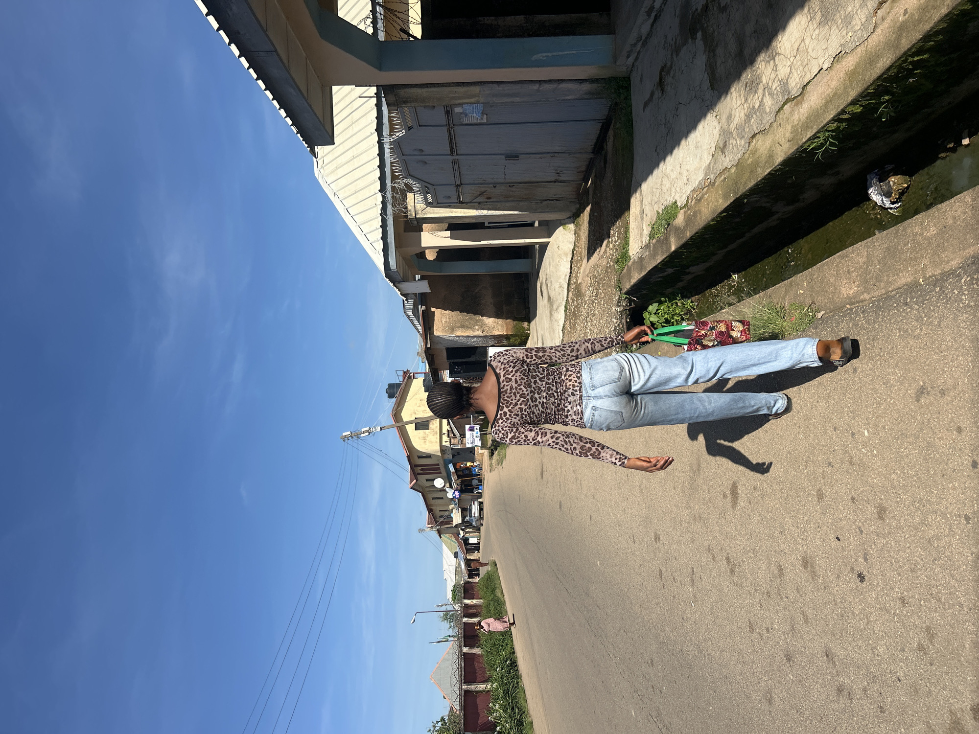 A young woman with dark braided hair, seen from behind, walks down an asphalt road in Jos, Nigeria. She wears a long-sleeved, leopard-print top and light blue jeans. In her right hand, she carries a floral patterned tote bag. The road is bordered by a concrete drainage ditch on the right, which contains murky water and some debris. The left side of the road features a row of buildings with corrugated metal roofs, fences, and some vegetation. A few other people and a car are visible in the distance along the road. The sky is a clear, bright blue with a few wispy clouds. The sun casts shadows, indicating it is daytime.