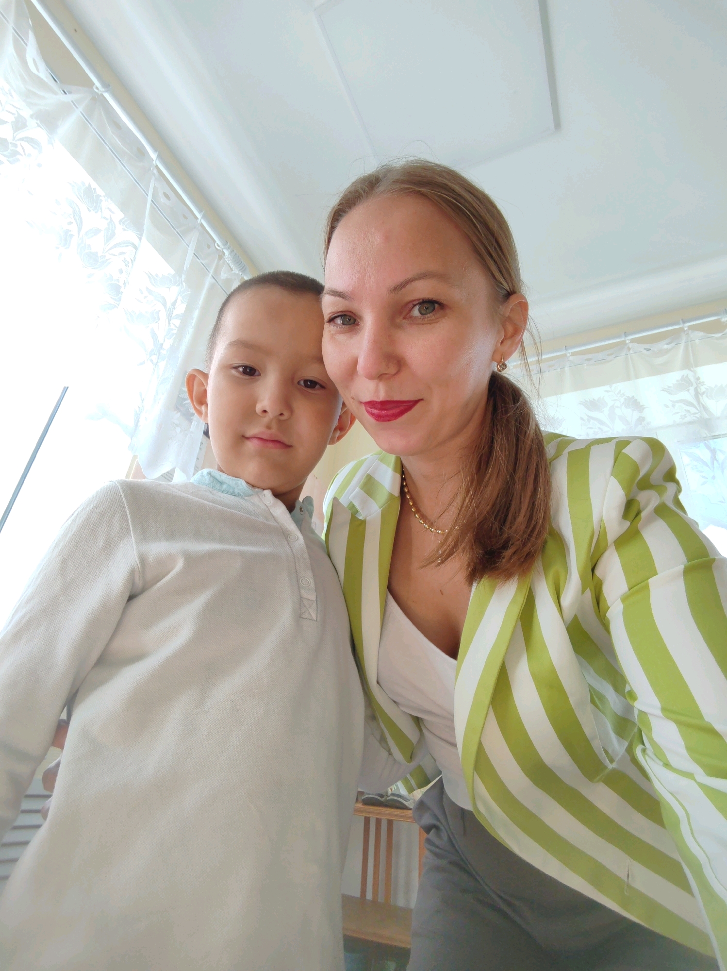 A woman and a child are photographed from a low angle in a room with bright, patterned curtains in the background. The woman, on the right, has her hair pulled back in a ponytail and is wearing a striped green and white jacket over a white top and grey pants. She is looking towards the camera with red lipstick. The child, on the left, has short dark hair and is wearing a white collared shirt. They are positioned closely together, with the child slightly in front of the woman. The overall lighting suggests an indoor setting during the day. The location is not specified as being in the city of Чухлэм, Russia.