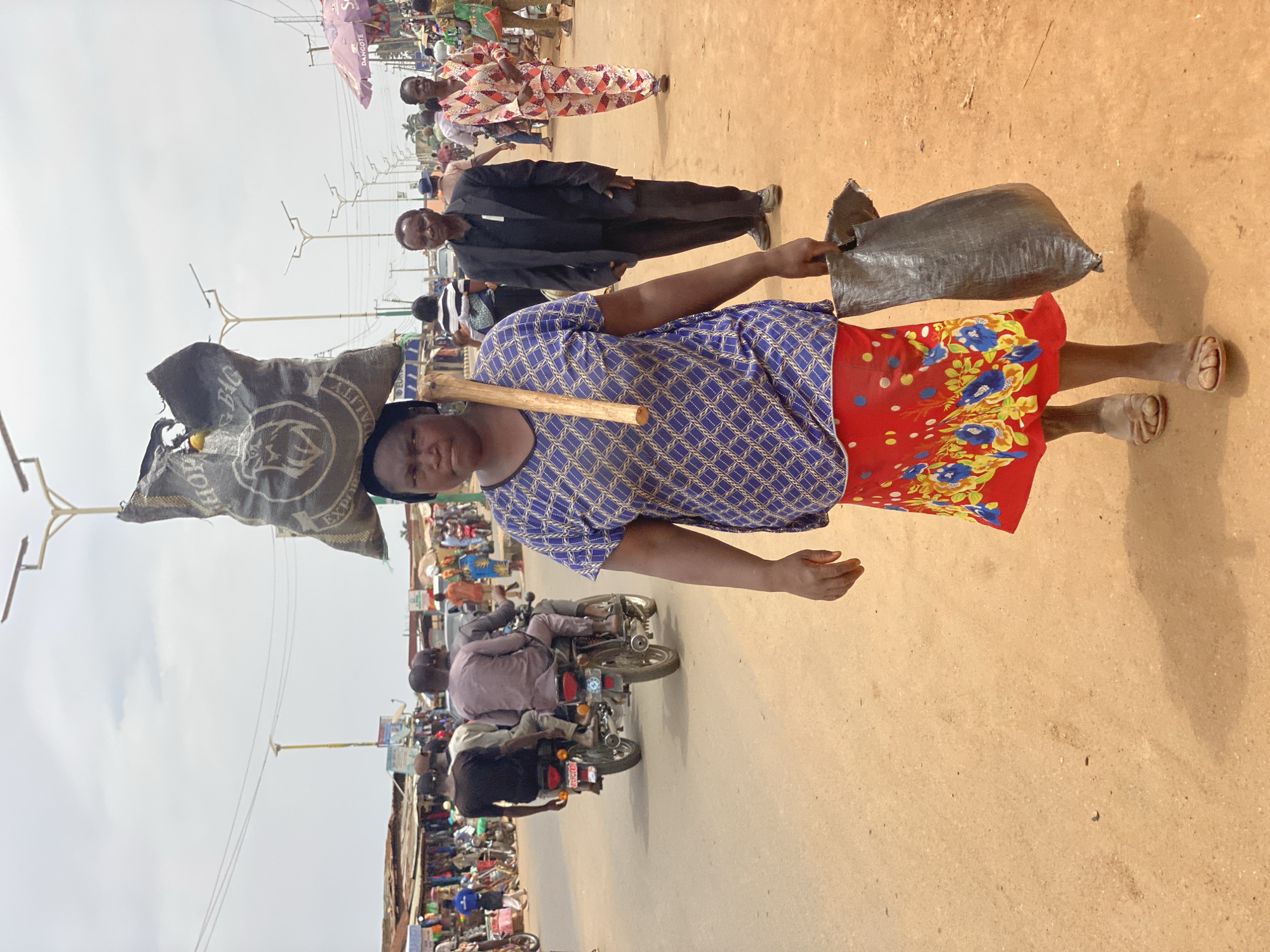 The image depicts a woman in Kafanchan B, Nigeria, carrying goods. She is wearing a blue patterned top and a red floral skirt, with sandals on her feet. She carries a sack on her head and a dark-colored bag in her hand. A wooden stick rests across her shoulders. In the background, there's a bustling street scene with pedestrians, motorcycles, and structures indicating a marketplace environment. The ground is reddish-brown, and power lines stretch overhead.