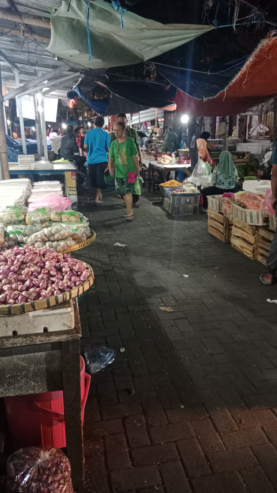 The image depicts an outdoor market scene at night, likely in Semarang, Indonesia, given the presence of a "Warung" sign and a price tag with "Rp." The market is covered by tarps of various colors, including blue, green, and orange, suspended from a metal frame. The ground is paved with dark bricks.
In the foreground, to the left, is a stall displaying a large quantity of red onions in a woven basket. To the right of the onions, there are bags of what appear to be garlic. Further back, on a table, are wrapped items, possibly food products, and in plastic bags.
Several people are visible in the market. A woman in a green shirt and patterned pants is walking through the aisle, carrying a pink bag. She is looking towards the right. Behind her, a man in a blue shirt and dark pants is also walking. Other individuals are partially visible or further in the background, some browsing stalls and others seemingly working.
The market appears to be active, with goods displayed for sale and people moving around. The lighting is a mix of overhead lights and possibly stall lighting, creating a somewhat dim but functional atmosphere. The overall impression is of a bustling night market.