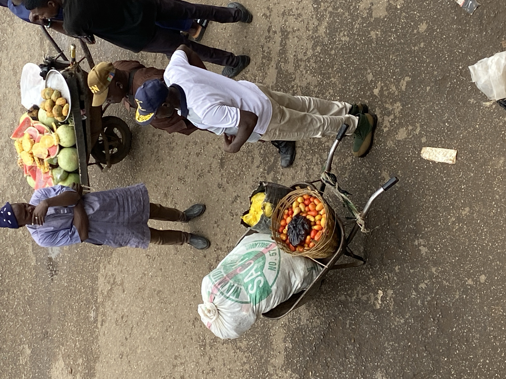This overhead shot depicts a street scene in Bukuru, Nigeria. Several individuals are present, alongside two carts used for selling goods.
One cart holds cut watermelons and other fruits. Three men stand near this cart, potentially interacting with the vendor.
The other cart, a wheelbarrow, is laden with a sack (labeled "SAC") and a basket filled with what appears to be dates or small plums. A man is standing near the wheelbarrow.
The ground is a light concrete surface. Other details include shoes, clothing, and miscellaneous discarded items on the ground.