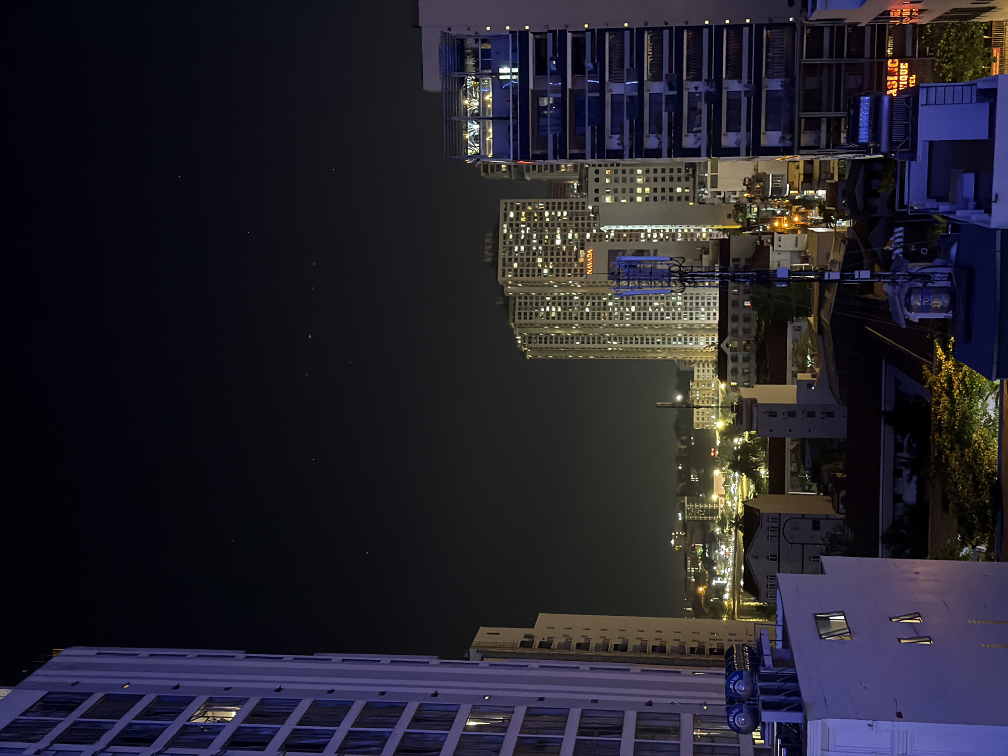 The image depicts a nighttime urban landscape in Bac Nha Trang Ward, Vietnam, viewed from an elevated perspective. The dark, clear sky shows several faint pinpricks of light, consistent with stars or distant celestial bodies.
In the immediate foreground, to the left, is the corner of a light-colored multi-story building, likely the vantage point, with one upper window emitting warm yellow light and three lower windows emitting cooler blue light. Directly below this, on a rooftop, are two cylindrical water tanks, one silver and one blue.
On the far left, a tall, white/light-grey high-rise building occupies the frame, featuring numerous rectangular windows, many of which are illuminated with warm yellow light. To its right and slightly further back, another similarly constructed light-colored building with multiple balconies is visible.
The central mid-ground is dominated by a cluster of several high-rise buildings, densely packed and extensively illuminated with yellow light from numerous windows. One prominent building displays a red sign reading "NAVADA." A tall, blue/grey communication tower, equipped with multiple antennas, rises from a lower building in this central area. Beneath the tower, an intricate network of smaller buildings, rooftops, and illuminated alleys creates a dense urban texture.
On the right side of the frame, a modern, dark-blue/grey multi-story building features balconies and large illuminated windows. A rooftop structure with railings is visible on its top. Partially visible signage on a building in the lower right reads "ASLIC" and "TIQUE," with "EL" further below, indicating "ASLIC BOUTIQUE HOTEL."
In the background, a discernible body of water (likely a bay or ocean) stretches across the horizon, bordered by a distant shoreline or promenade illuminated by a line of artificial lights. Faint outlines of hills or mountains are visible beyond the illuminated urban sprawl. Artificial lighting from buildings, streets, and signs is pervasive, casting a glow over the city. No people are discernible in the image.