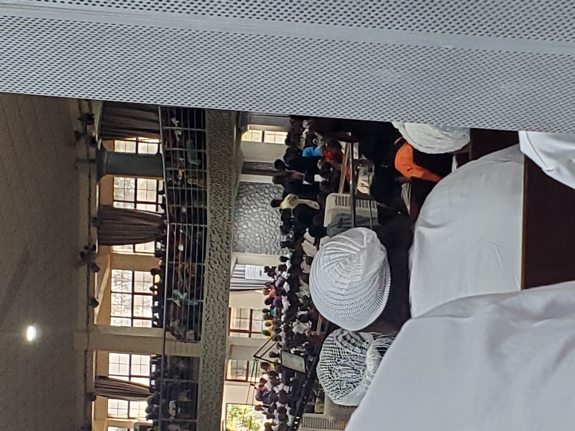 This media file shows a gathering of Muslim men inside what appears to be a mosque in Kason, Nigeria. The image is taken from a high angle, looking down on the congregants. The men are dressed in traditional attire, primarily white robes and caps, and are engaged in prayer. Some are standing, while others are in a kneeling or prostrating position, indicating the prayer is in progress.
The setting features architectural elements typical of a mosque, including windows with decorative grilles and textured stone walls. The lighting suggests it could be daytime, possibly late afternoon or early evening, as there is natural light filtering in from the windows. The overall atmosphere appears solemn and focused, consistent with a religious service. The presence of multiple rows of people praying together signifies a communal prayer session.
Notable details include the variety of kufi caps worn by the men, ranging from simple white ones to more intricately patterned designs. There is a sense of community and shared devotion among the individuals. The image is a snapshot of a religious event, capturing a moment of collective worship. No visible text is present in the image.