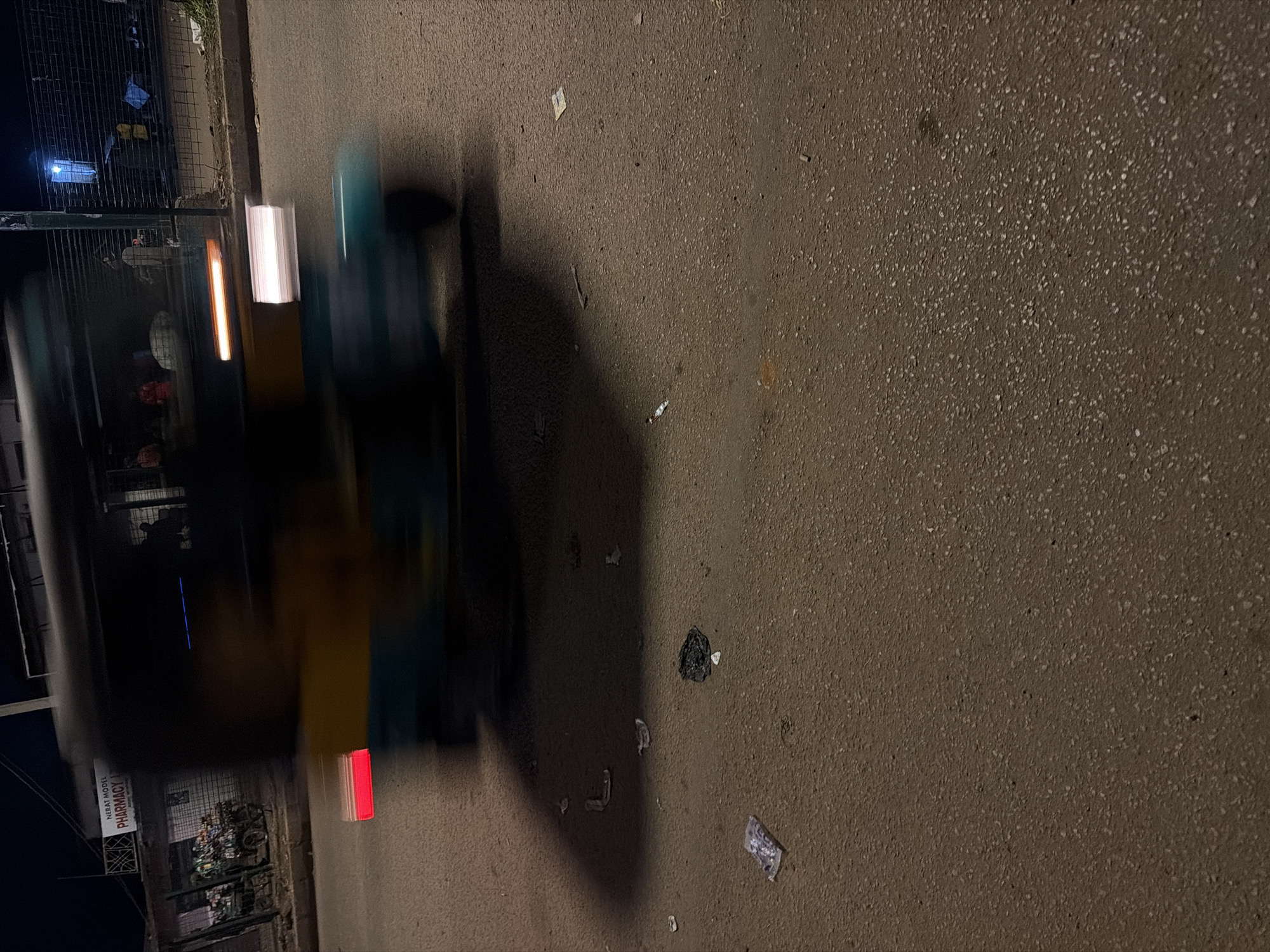 The image depicts a nighttime scene on a street in Bukuru, Nigeria. A blurred auto-rickshaw is the primary subject, in motion, obscuring details of its passengers and specific features. Red and white lights are visible on the vehicle, suggesting headlights and taillights. The street itself is paved and appears to have some debris scattered on its surface, including what looks like paper or plastic wrappers and a dark, crumpled object. In the background, a storefront with a sign reading "NERAT MODEL PHARMACY" is partially visible, along with some stacked items and a wire fence. The overall lighting suggests a low-light environment, possibly streetlights or the vehicle's own illumination.