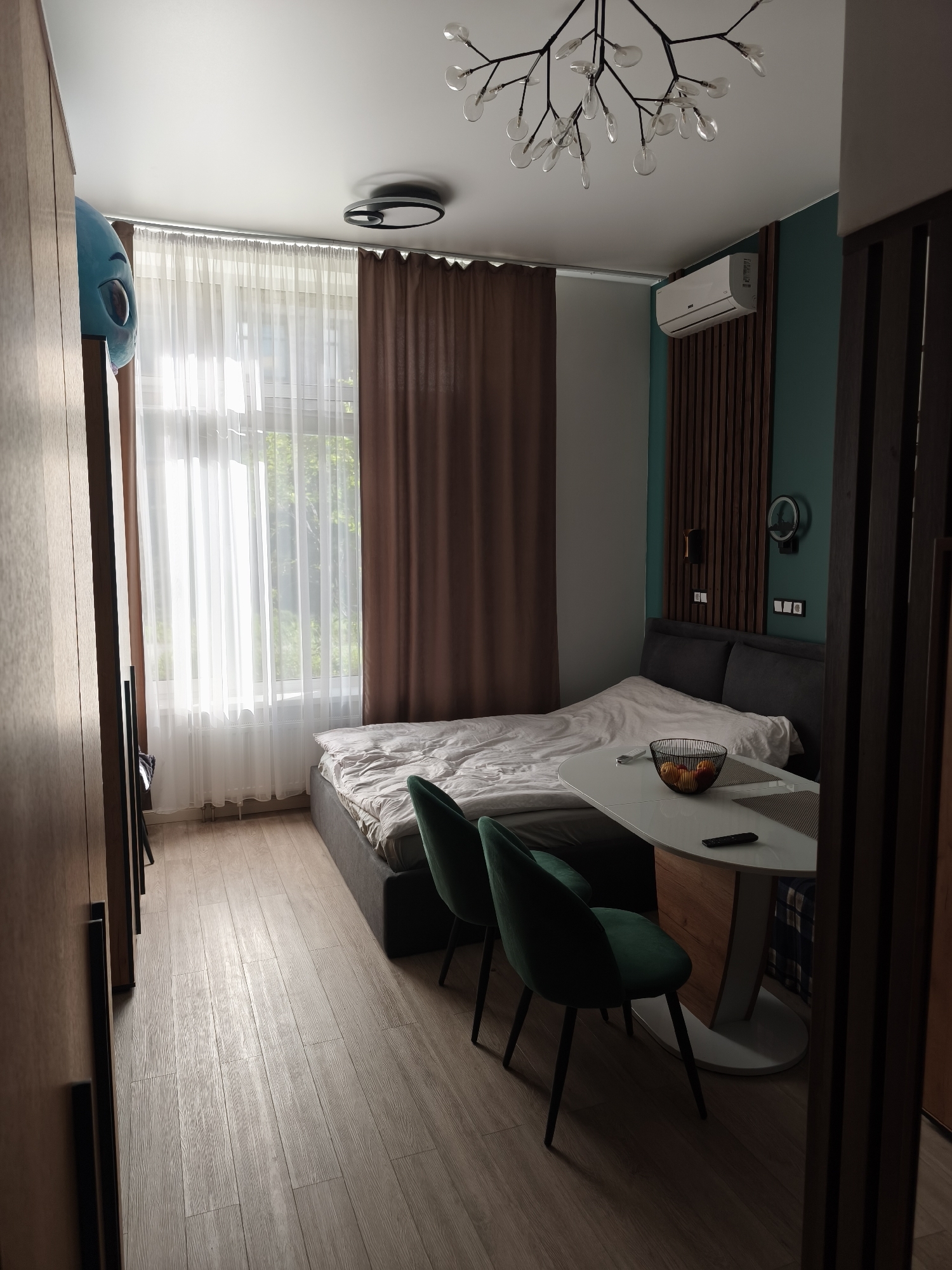 The image shows an interior view of a combined living space and bedroom. Dominant features include a bed, a dining table with two green chairs, and a large wardrobe. The bed is grey, with rumpled white bedding. The dining table is white with a wooden base, and a black wire basket containing fruit, along with a remote control, rests on its surface. The chairs are upholstered in a dark green fabric with black legs. A wardrobe is located on the left side of the image. The window is covered by sheer white curtains and heavier brown drapes. An air conditioning unit is mounted on the wall above the bed, which has a dark green accent wall behind it featuring vertical wooden slats. A modern chandelier hangs from the white ceiling. The floor is covered in light wood-look planks. There is no explicit indication of the location in Moscow, Russia.