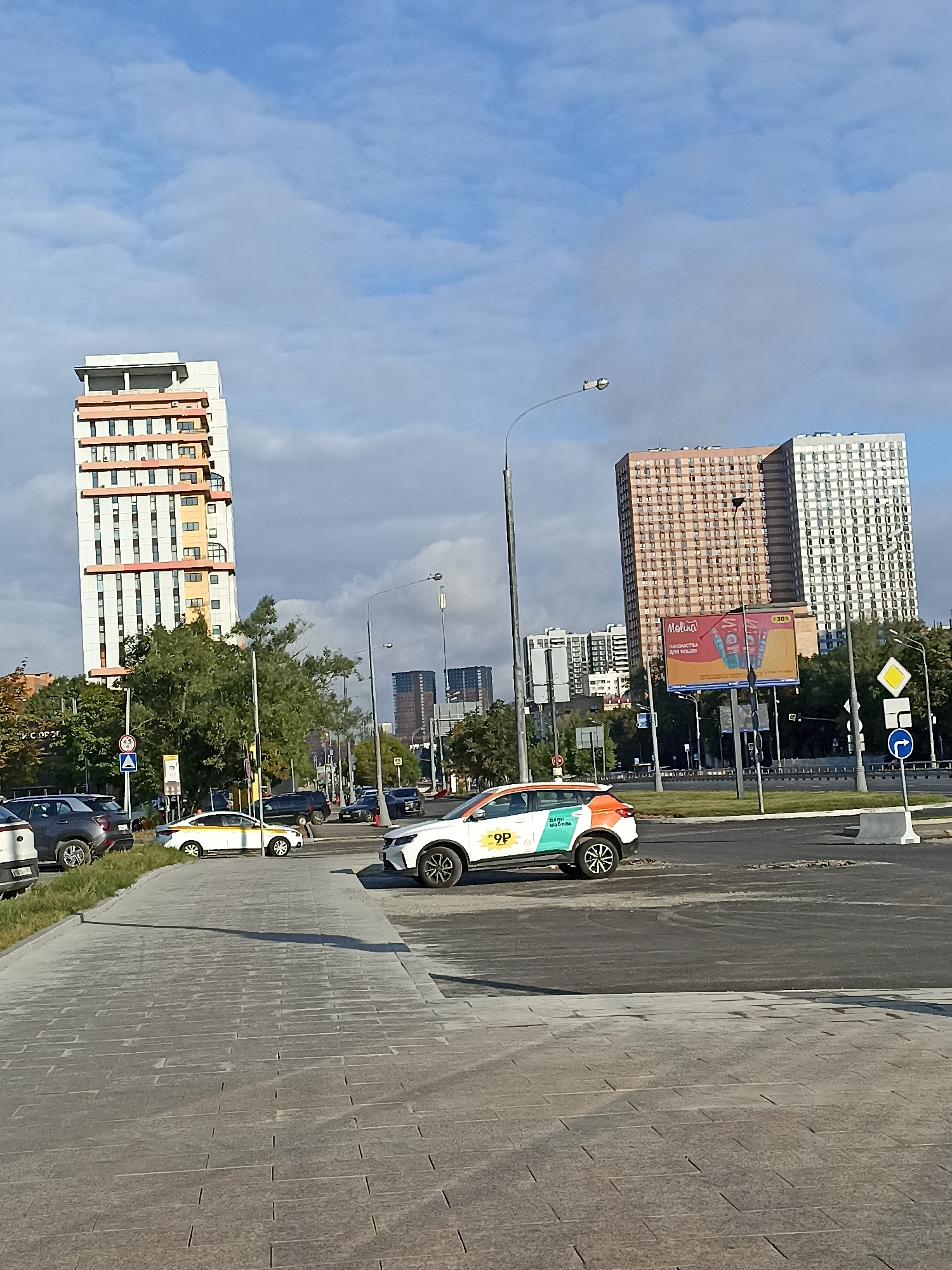 The image captures a street scene in Moscow, Russia, on a partly cloudy day. In the foreground, a paved pedestrian walkway runs diagonally from the bottom left to the middle right of the frame. To the right of the walkway is a section of asphalt road, showing some wear and tear. A white car with colorful branding, including the numbers "9P" and Russian text, is parked on the right side of the road. Further down the road, a white taxi with a yellow stripe is visible.
In the midground, several cars are driving on the road. A tall, slender building with white and orange sections stands prominently on the left side of the image, partially obscured by trees. Behind it, a line of green trees creates a natural boundary. Further in the background, several tall residential buildings are visible. Two large apartment buildings, one with a beige facade and the other with a white facade, are particularly noticeable. A billboard with advertisements is positioned between these buildings. The sky above is blue with scattered clouds. Streetlights and traffic signs are also present along the road.