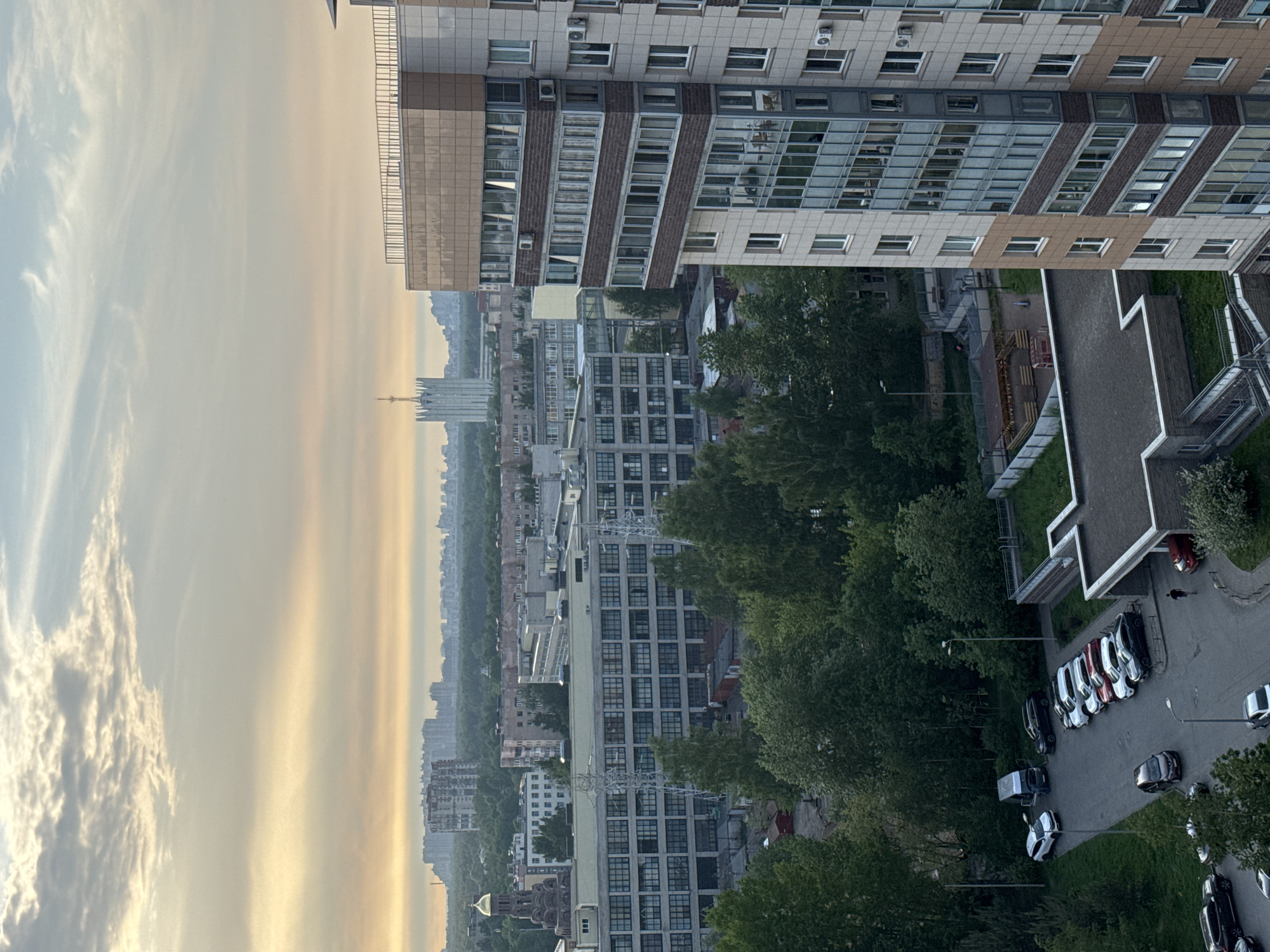 The image depicts an urban landscape in Unknown, Russia, with a mixture of high-rise apartment buildings and older structures. A modern high-rise occupies the top portion of the image, showcasing a grid of windows and architectural details. Below, trees are seen lining the streets. Further in the distance, the skyline is punctuated by a prominent spire. At the bottom right of the image, cars parked on the side of the road are visible. The sky is a mix of clouds and a hazy orange glow, suggesting either sunrise or sunset.