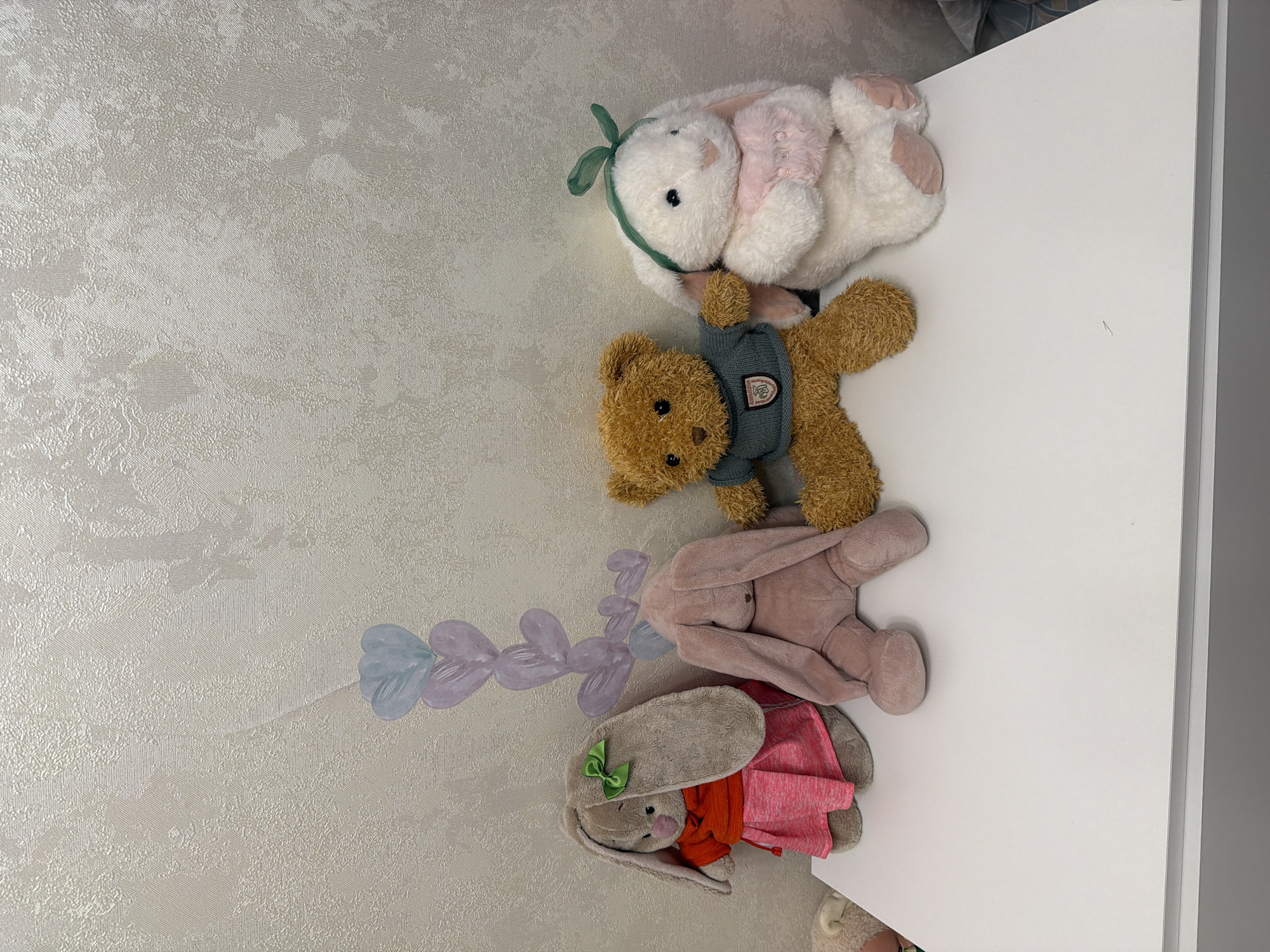 Four stuffed toys, three rabbits and one teddy bear, are arranged on a white surface against a textured, light-colored wall. From left to right, the first stuffed toy is a light brown rabbit wearing a pink skirt and an orange top, with a green bow on its head. Next to it is a dusty rose colored rabbit in a sitting position, with its head bowed. To its right is a brown teddy bear wearing a blue sweater with a crest. The fourth stuffed toy is a white rabbit with pink accents, wearing a pink scarf and a green bow on its head. Behind the toys, on the wall, is a pastel drawing of a vertical chain of hearts in shades of purple and blue. The white surface appears to be a shelf or the top of a piece of furniture. The lighting is even, suggesting an indoor setting.