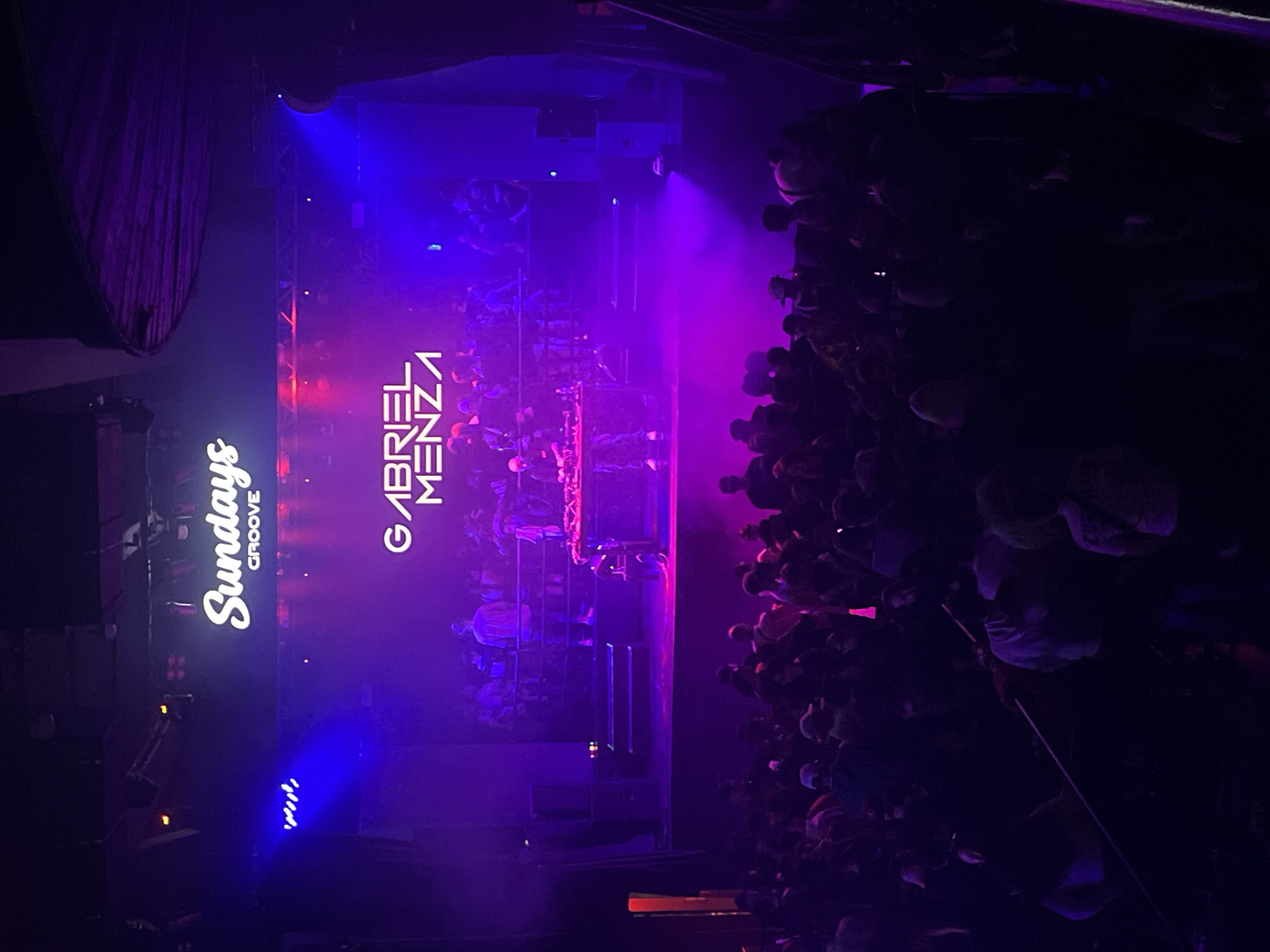 A vertical, high-angle shot shows a crowded event stage at "Sundays Groove" in Buenos Aires, Argentina. The stage displays "GABRIEL MENZA" vertically in white letters. Above the stage, a lighting rig casts blue and purple light. A large crowd is tightly packed to the right of the stage. There are dark curtains on the left.