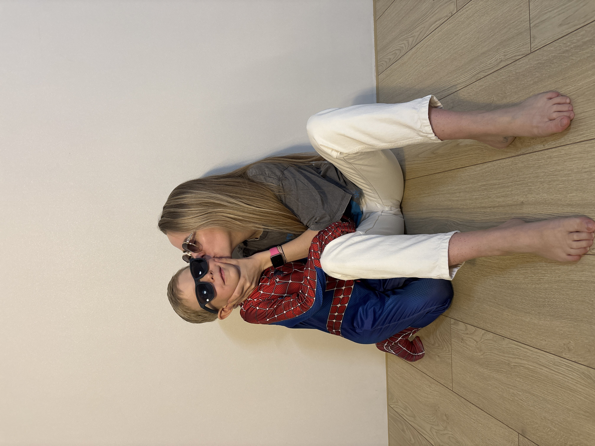 The image shows a room with a white wall and a light brown wood floor. A woman with long blonde hair and sunglasses is kissing a young boy wearing a Spiderman costume and sunglasses. They are both sitting on the floor, angled against the wall. The woman is wearing white pants and a gray t-shirt. The boy's Spiderman costume is predominantly blue and red. The location is unclear, and there is no evidence to indicate the image was taken in Chaikovsky, Russia.