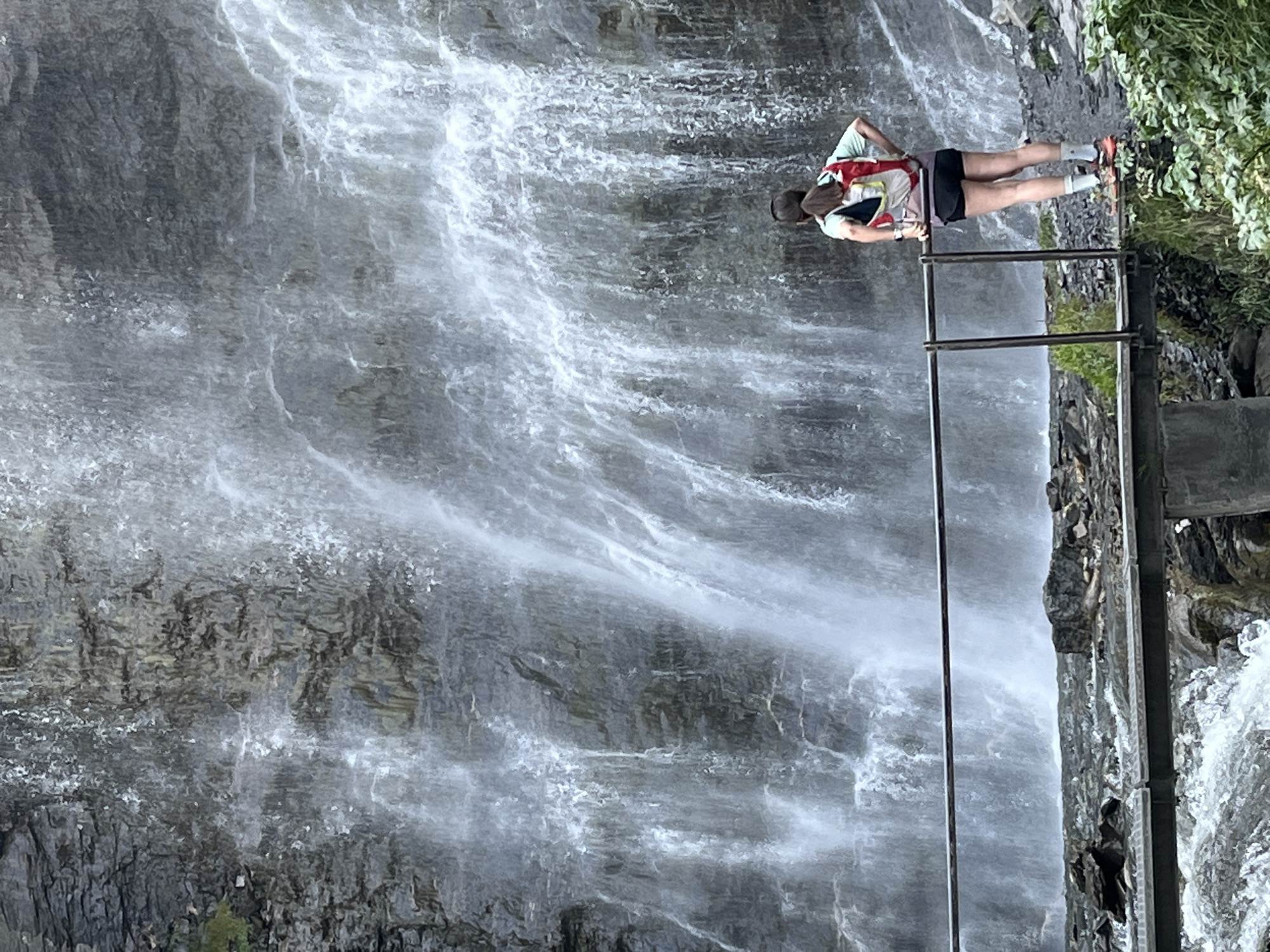 A person stands on a metal bridge, facing a tall waterfall that cascades down a rocky cliff. The person, viewed from behind, wears a light green shirt, black shorts, white socks, and athletic shoes. They have a backpack with red, white, and yellow accents. A metal railing runs along the bridge and the person's right hand rests on it. The waterfall is the dominant feature, with white water flowing over grey rock. The ground around the base of the waterfall is rocky, with some green vegetation visible to the right. The water below the bridge is turbulent and white, indicating a fast current. The scene appears to be an outdoor natural setting. The image is likely taken in the vicinity of Benasque, Spain, given the presence of a waterfall and mountainous terrain common to the region.