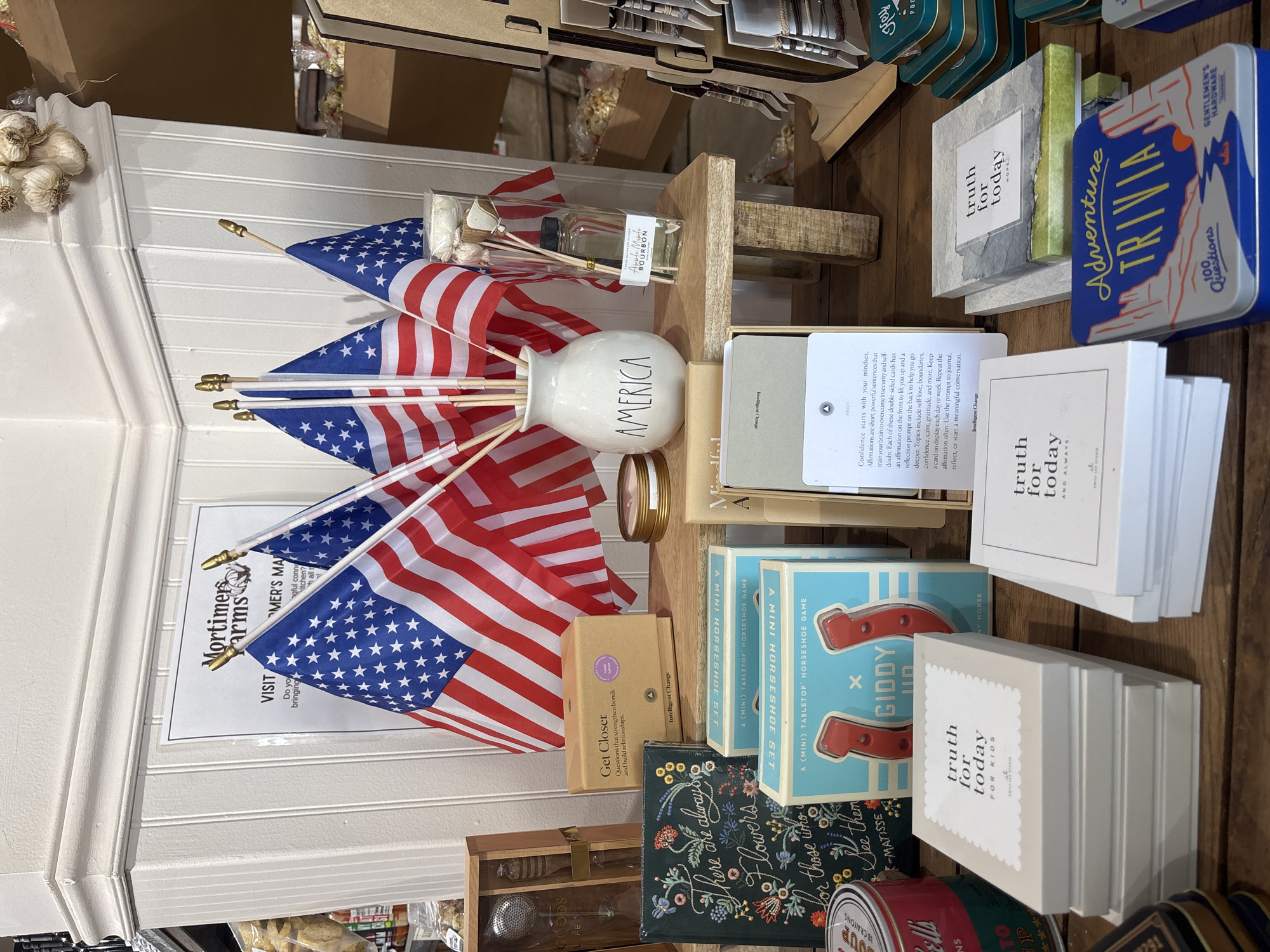 The image depicts a retail display in Prescott Valley, Arizona. The display features a collection of merchandise arranged on wooden shelves and a rustic wooden stool. Prominently displayed are four small American flags in a white vase labeled "AMERICA." To the left of the flags, a box for a "Get Closer" game is visible, along with a dark green book with floral embroidery that reads "There are always Flowers for those who see them." Below these items, two boxes for a "Giddy Up A (Mini) Tabletop Horseshoe Game" are stacked.
Further to the right, on the stool, is a glass diffuser with reeds, labeled "Amber Mountain Candle Bourbon." A small round candle tin is also present. In front of the stool, to the left, are stacks of beige boxes labeled "truth for today for kids." To the right of these, on the wooden surface, is a box titled "truth for today HOPE." Several rectangular tin boxes with various designs are visible in the foreground and right side of the image, including one labeled "Adventure TRIVIA." A partial view of another tin box with red and white lettering is visible at the bottom left. The background consists of a white paneled wall with a framed sign that reads "Mortimers Farms." Garlic bulbs are visible hanging from the top of the wall. The overall setting appears to be a gift shop or a similar retail environment.