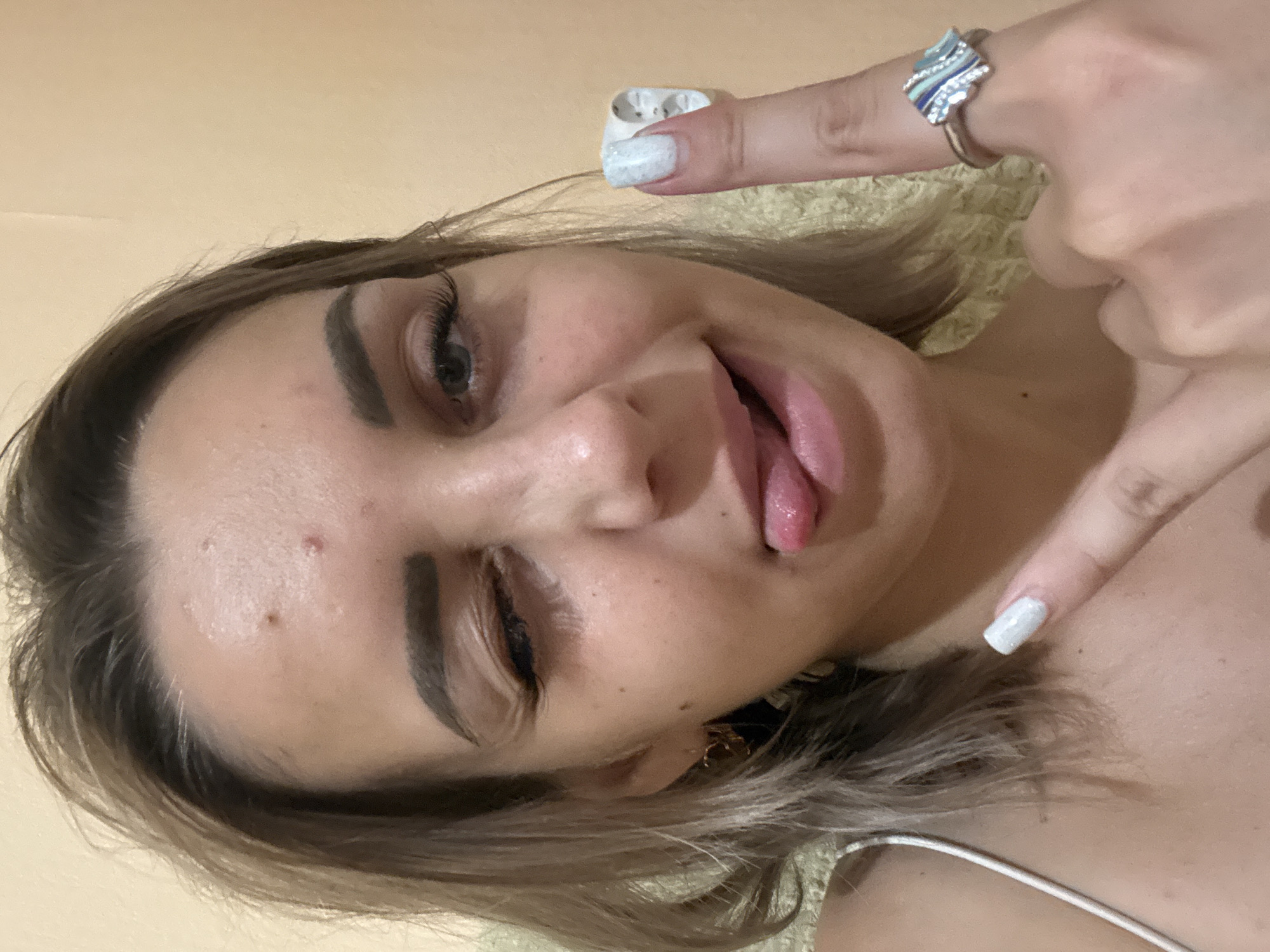 The image is a close-up, self-taken photograph of a young Caucasian woman with blonde hair and fair skin. She is winking with her left eye and sticking her tongue out. She is wearing a white tank top. On her right index finger, she has a white-patterned nail polish and is wearing a silver ring with blue and white stones. Her right thumb is also visible, and it appears she may be holding a small, white object, possibly a piece of paper or cotton. Her skin shows some minor blemishes on her forehead. The background is a plain, light-colored wall, and some textured yellow fabric is visible behind her. There is no indication of the city of Kholmsk, Russia, within the image itself.