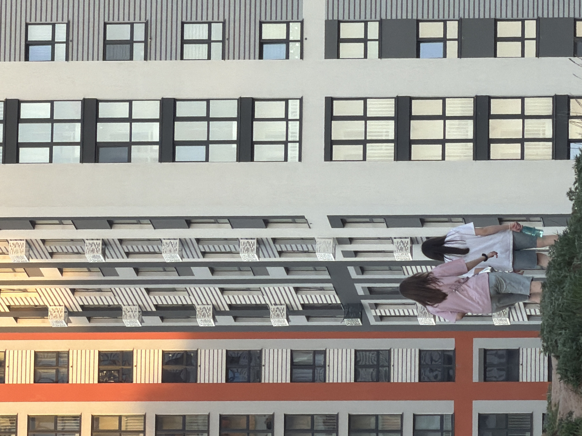 Here is a detailed description of the image:
**Scene:** The image depicts an exterior shot of a multi-story building in Izhevsk, Russia. The building has a modern architectural style, with distinct horizontal layers. There are two women in the foreground in the building's reflection in a window.
**Building Details:**
* The building has several floors, each with rows of windows.
* The window styles and sizes appear to vary slightly between floors.
* One section of the building has an orange stripe with a cross design, while others have striped or geometric designs.
* There are decorative elements or awnings above some of the windows.
**People:**
* Two women are partially visible in the reflection in one of the building's windows.
* One is wearing a pink top and gray shorts, and the other is wearing a white top and gray shorts.
* Their interactions are unclear.
**Environment:**
* Some foliage (likely trees or bushes) is visible to the right of the building.
* The weather appears to be clear, allowing for a clear reflection in the window.