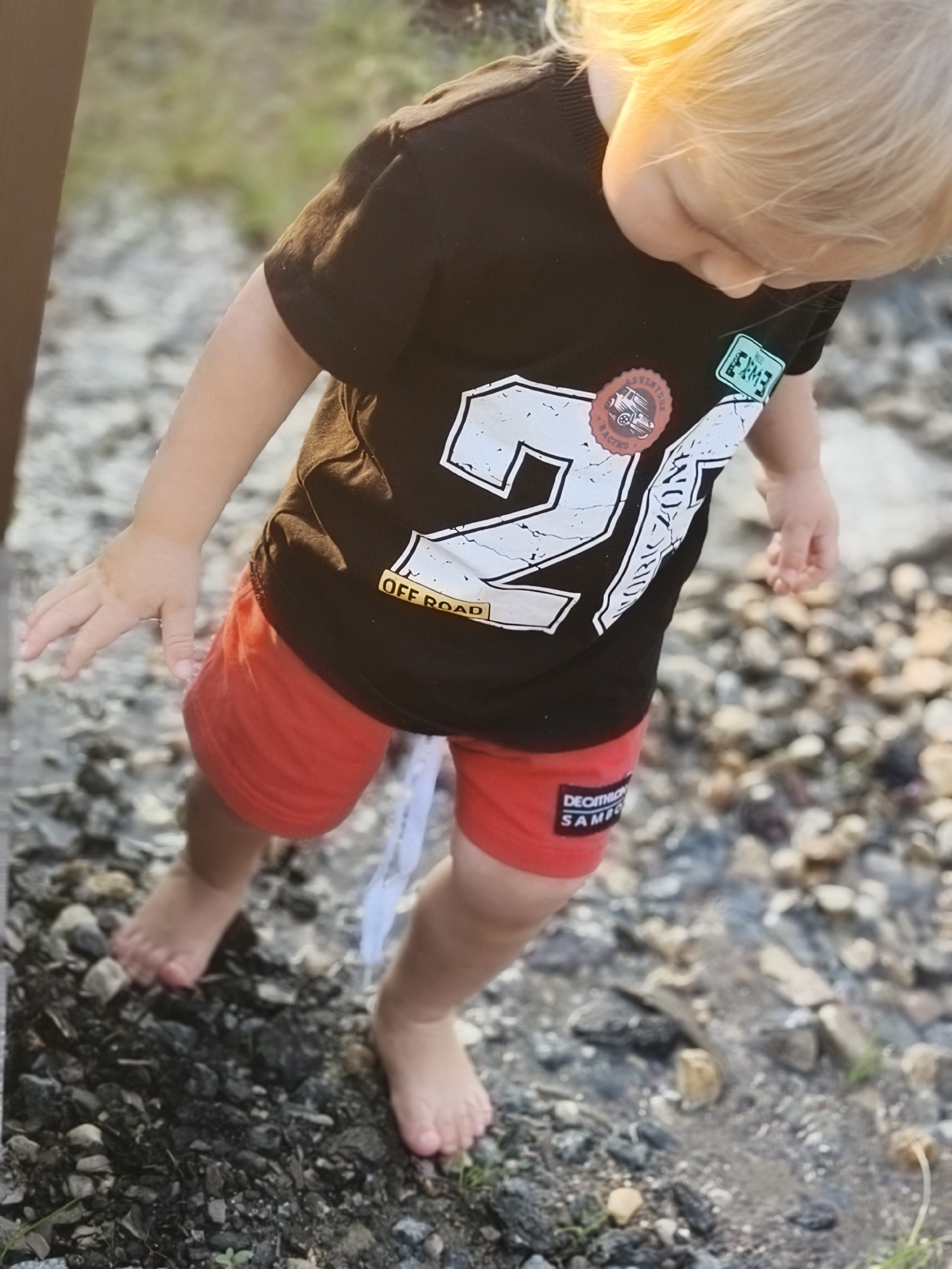 A young child or toddler with blonde hair, illuminated by warm sunlight, is depicted from an elevated perspective, looking down towards the ground. The child wears a dark (likely black or dark brown) short-sleeved t-shirt featuring prominent white graphics, including a large '27' (or similar numerals), the text 'OFF ROAD,' and two circular badges. One badge shows an illustration of a vintage vehicle with 'ADVENTURE RACING' text; the other is a light green rectangular patch with faint white text/numbers such as 'RXM3'. The child also wears red shorts, which have a white label on the left leg displaying 'DECATHLON SAMC' (or similar branding). Both feet are bare, and the child's left hand is slightly extended forward. A thin, white, strip-like object hangs from the child's right hand or arm towards the ground. The child stands on uneven terrain composed of various sizes of dark and light rocks, gravel, and soil, with sparse green vegetation visible. A damp or wet patch of ground is present to the child's right. The background is blurred with soft green and brown tones, suggesting an outdoor environment. The scene takes place in the city of Unknown, Unknown.