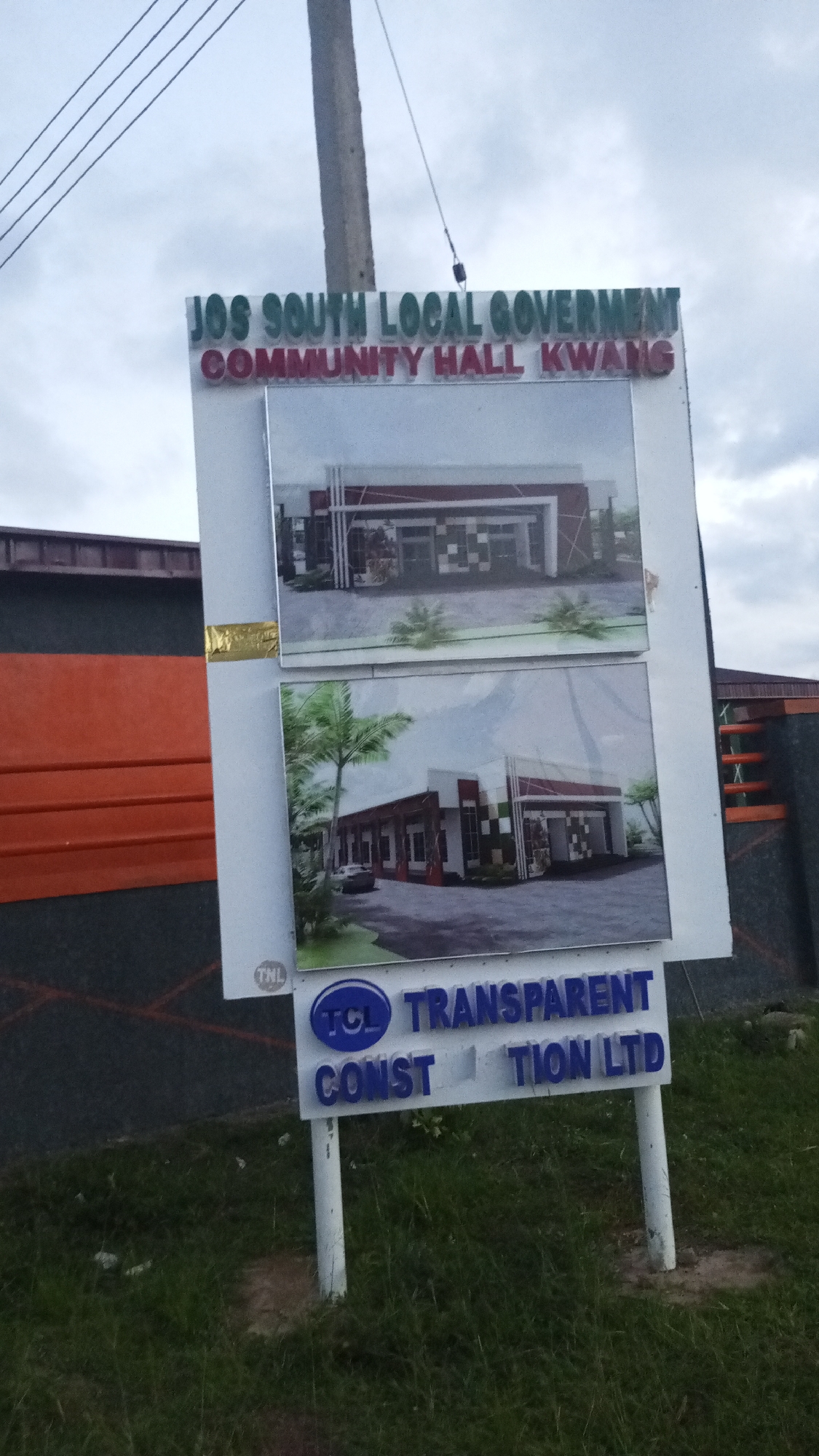 This is a photograph of a sign for the Jos South Local Government Community Hall Kwang in Nigeria. The sign is white with blue and green lettering and features two architectural renderings of the community hall. The renderings show a modern building with a red and white facade. The sign is supported by two white posts and is located in an outdoor setting with green grass and a cloudy sky. In the background, there is a building with orange and dark grey siding.