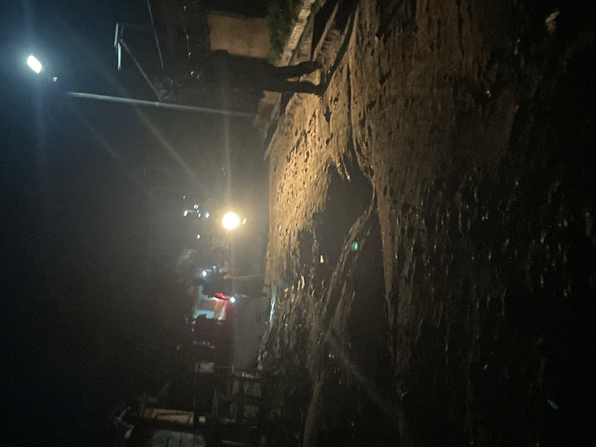 The image depicts a nighttime scene on a dirt road in Lokoja, Nigeria. The road is uneven with visible puddles and debris. A street light illuminates a portion of the scene, casting long shadows. In the foreground, a person walks away from the camera, silhouetted against the light. Further down the road, other individuals and possibly a vehicle with illuminated headlights are present. Structures, likely houses or shops, are dimly visible on either side of the road, partially obscured by the darkness. The overall impression is of a rural or semi-urban environment at night, with limited artificial lighting.