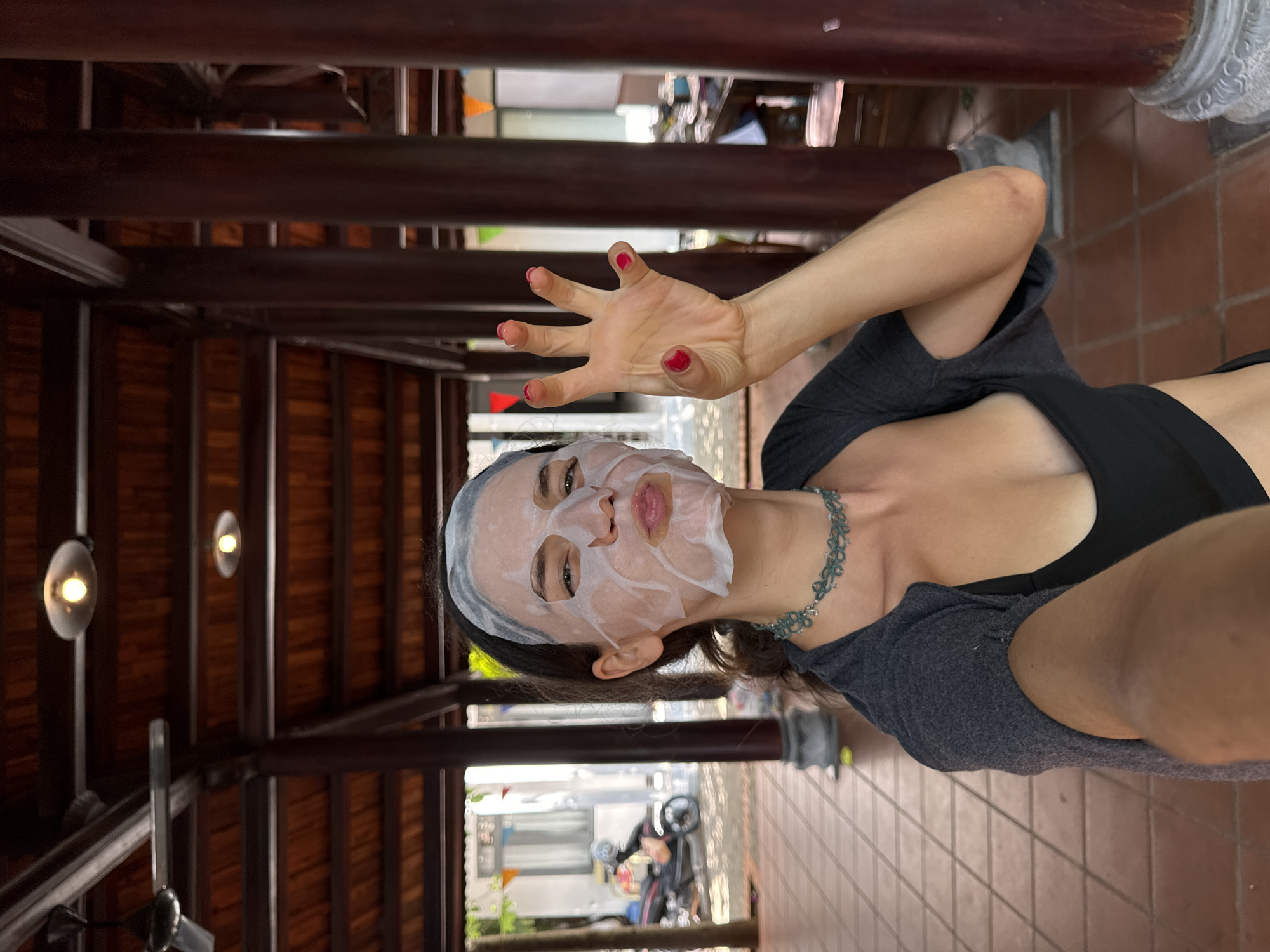 In Mũi Né, Vietnam, a woman is taking a selfie in a covered outdoor area with a tiled floor and dark wooden pillars and ceiling. She is wearing a black tank top, a grey cardigan draped over her shoulders, and a face mask. Her hand, with bright red nail polish, is raised and her fingers are splayed. In the background, a scooter is parked near buildings.