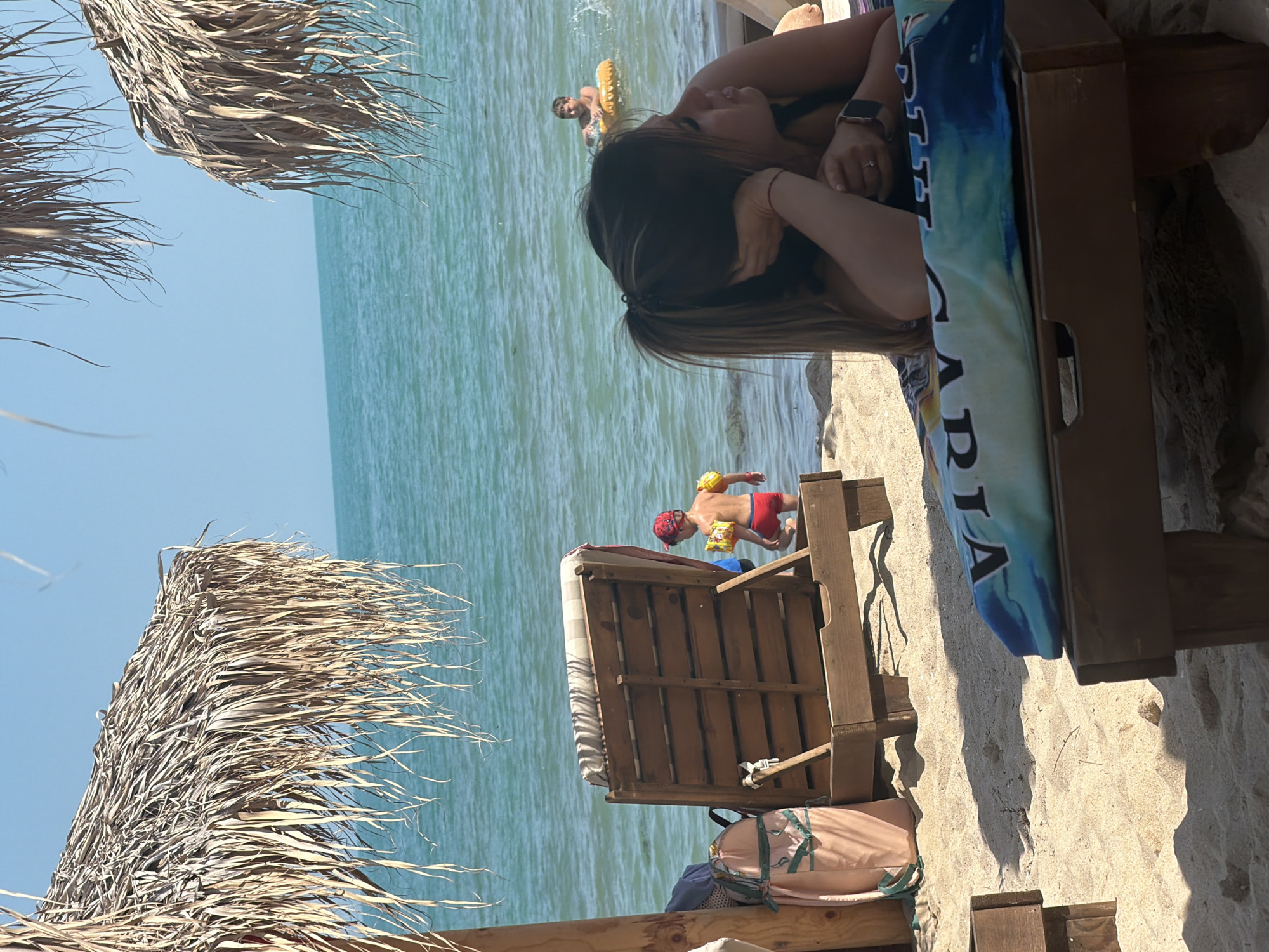 This image depicts a sunny beach scene in Kranevo, Bulgaria. The sky is clear and bright blue, meeting a tranquil, turquoise-colored sea on the horizon. The foreground and midground consist of light-colored sand, showing shadows cast by beach equipment under a high sun.
In the upper left, a large straw beach umbrella is partially visible, providing shade. Another straw umbrella is partially visible in the upper right.
On the left side of the midground, a wooden beach chair or lounger with a striped (grey/white) cushion is positioned facing the sea. A peach or pink-colored beach bag or soft item leans against the lounger's leg.
In the lower right foreground, a woman is lying prone on a wooden beach lounger, covered by a blue, white, and black patterned cushion or towel with partial text visible ("VIAG..."). She has long, dark hair tied in a ponytail and is wearing a dark swimsuit top. Her left arm is bent, supporting her head, while her right hand rests near her left arm. A ring is visible on her left ring finger, and a dark-strapped wristwatch is on her right wrist. Her gaze is directed slightly downwards or towards the right.
Two children are in the sea. In the mid-ground, slightly to the left, a young child, possibly a toddler, wearing red swimming trunks, yellow arm floats, and a red cap, is walking into the shallow water with their back to the camera. Further out in the water, to the right of the first child, an older child is floating in a yellow, ribbed inflatable ring, facing towards the left, with water splashing around them.