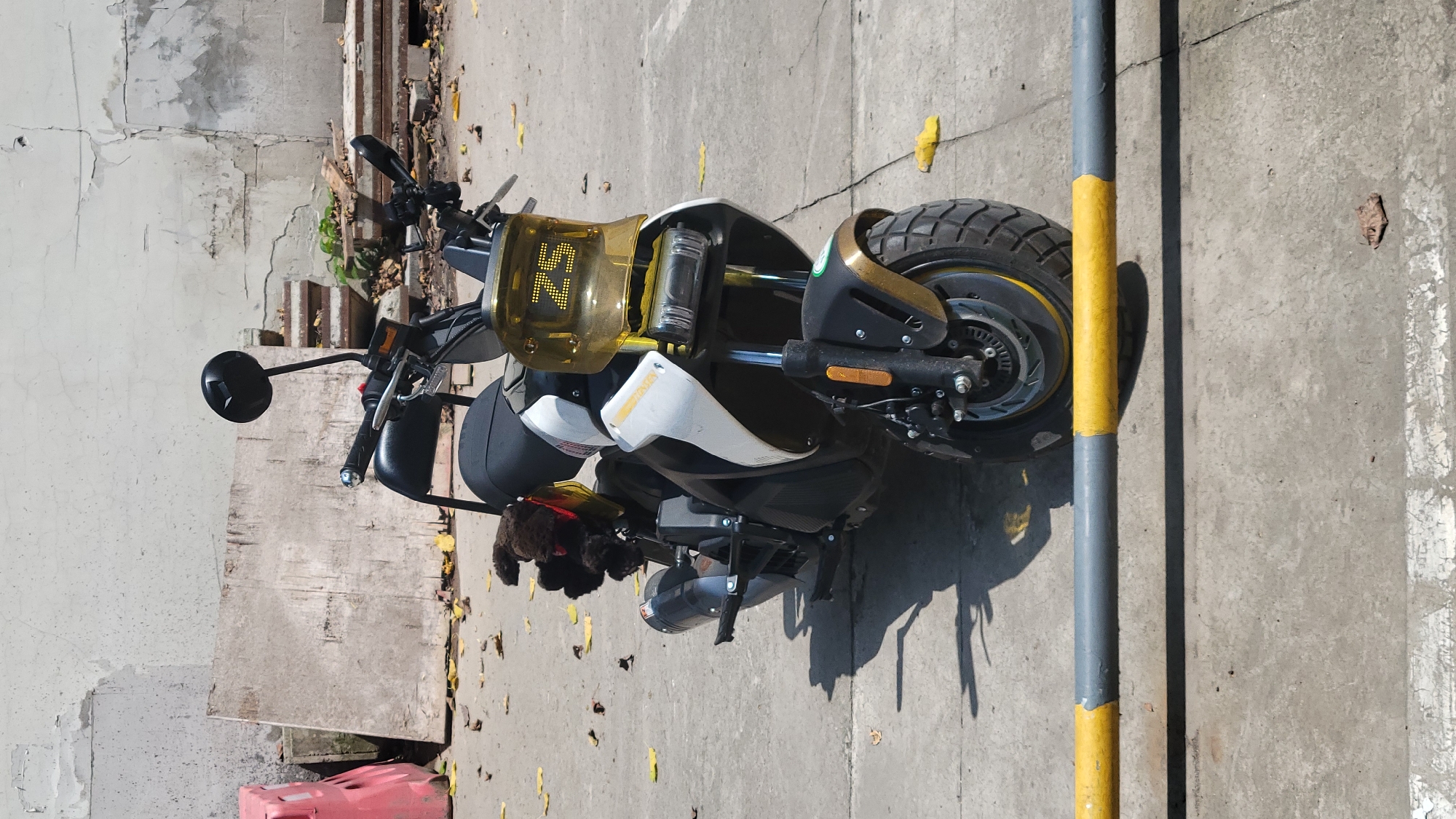 The image, captured in Yuzhong District, China, shows a modern electric scooter or motorcycle parked outdoors on a concrete surface during a bright, sunny day. The vehicle features a sleek design with a white and black body, complemented by gold or bronze accents on its fairings and wheel. It is equipped with large, knobby tires, suggesting off-road capability.
A prominent transparent yellow-amber windscreen on the front of the scooter displays digital text: "25" at the top and an "N" below it, likely indicating speed and neutral gear. Black handlebars with circular mirrors extend from the front, with a visible red brake lever. A small, dark, furry object, possibly a toy or a glove, is tucked near the handlebars. The scooter casts a distinct shadow on the ground, confirming the clear and sunny weather.
The ground is light gray concrete pavement, showing various cracks and scattered yellow leaves. To the right of the scooter, a gray and yellow striped bollard stands, casting a long shadow. In the background on the left, a worn, light-colored wall with extensive cracking and peeling paint or plasterwork is visible. Below the wall, there are some rough concrete steps or debris, along with a small patch of green plant life. A low-lying red plastic object is also partially visible in the bottom-left corner of the frame. No people are present, and the scooter appears stationary.