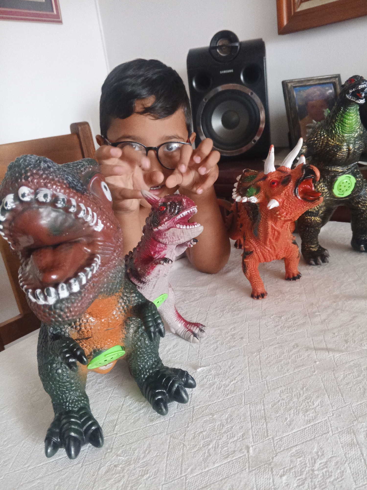 This indoor photograph captures a playful moment, likely in a home in Caracas, Venezuela, centered around a young boy and his collection of dinosaur toys. A boy with dark hair and glasses is partially visible in the mid-ground, peeking over and between the toys. His hands are raised near his face, suggesting active engagement with the dinosaurs, perhaps mimicking them or making them "roar."
Four distinct dinosaur toys are arranged on a white, textured surface, possibly a tablecloth. In the left foreground, a large, green and orange dinosaur, resembling a Tyrannosaurus Rex, commands attention with its wide-open red mouth full of white teeth and fierce expression. Adjacent to it is a smaller, slender dinosaur with a pink and red coloration, also featuring red eyes and an open mouth.
Towards the right, an orange Triceratops-like dinosaur stands prominently, characterized by its white horns, red eyes, and open maw. Behind it, a green and gold dinosaur, possibly a Godzilla figure, stands with red eyes and an open mouth, distinguished by a circular green element on its torso.
The background elements include a wooden chair back on the left and a black Samsung speaker on the right, which bears the visible text "SAMSUNG." Next to the speaker, a framed picture is partially seen. The overall lighting suggests it is daytime. The image conveys a sense of childhood imagination and the excitement of playing with toys.