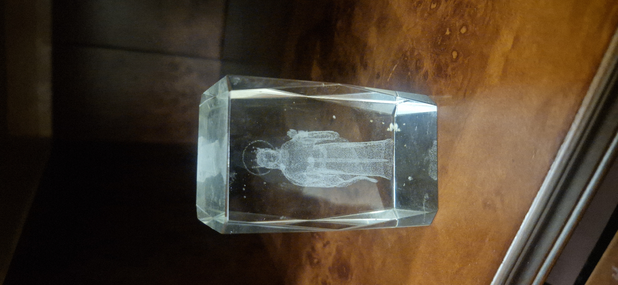 The image shows a clear, rectangular glass or crystal block lying on a wooden surface. An image of a person with a halo is etched or embedded inside the block. The person appears to be wearing robes or garments. The wooden surface has varying shades of brown. The environment appears indoors. The city is Unknown, Unknown.