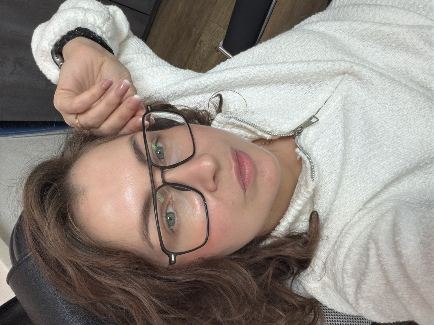 An eye-level close-up shows a woman with fair skin, green eyes, and long, wavy brown hair, lying down and looking directly at the camera. She is wearing black-framed glasses that highlight her eyes. Her expression is neutral and calm, with slightly parted pink lips.
She is dressed in a white, textured, possibly fleece or boucle material jacket or hoodie, partially unzipped with a silver zipper pull visible near her neck. A delicate silver chain necklace can be seen around her neck. Her left hand is resting near her head, showcasing a thin gold ring on her ring finger and a black braided band, possibly a watch strap, on her wrist. Her nails have a natural French manicure style.
The background is simple and suggests an indoor setting. Behind her head and left arm is a dark, possibly black, upholstered surface, resembling a chair or headrest. A wooden floor is visible in the upper right portion of the image. The lighting is soft and even, indicating an indoor environment. There is no visible text. The overall scene conveys a casual and relaxed moment, possibly a selfie taken indoors in Chaikovsky, Russia.