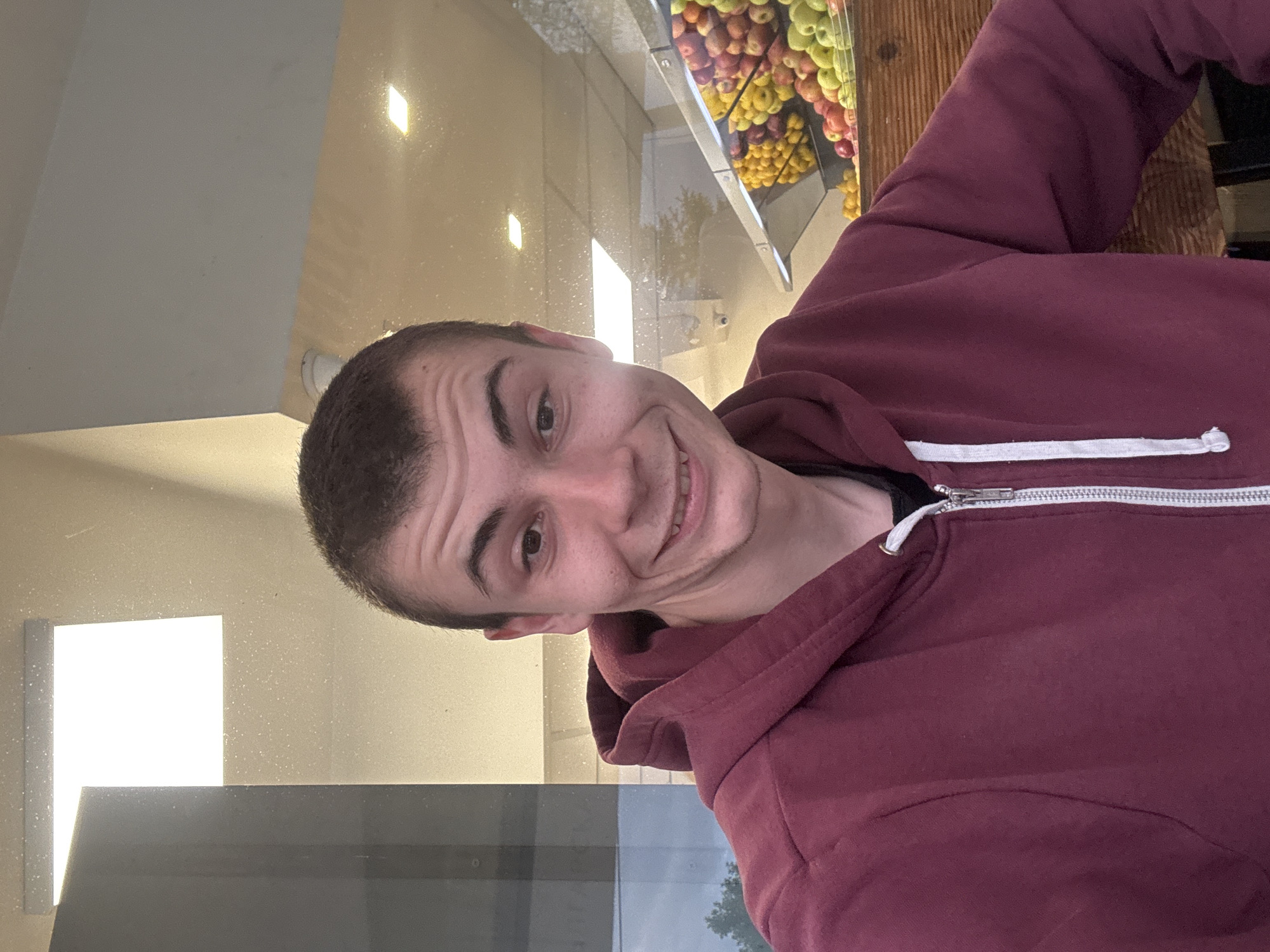 A man wearing a maroon hoodie is visible in the foreground. The background appears to be a store window, reflecting the outside. Visible through the window is an assortment of apples. The location is most likely in Plovdiv, Bulgaria.