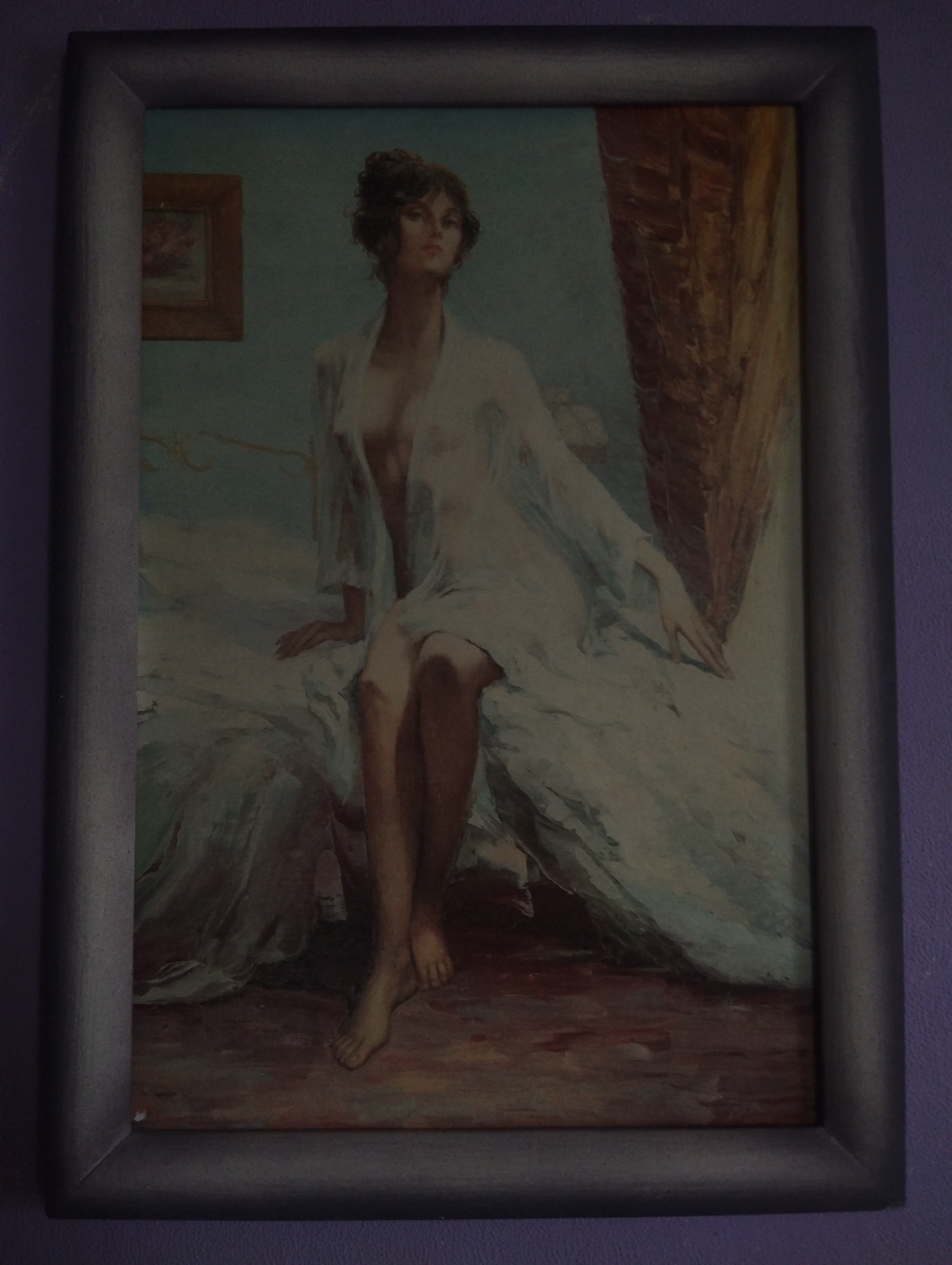 A framed painting depicts a woman sitting on a bed. The woman has dark hair styled up and is wearing a sheer white robe. Her left arm is bent and resting on her lap, while her right arm is extended slightly forward. Her legs are crossed at the ankles. The bed appears to be covered in white linens. In the background, to the left of the woman, is a small framed picture on the wall, and to the right is a richly textured brown curtain or drapery. The overall color palette is muted, with blues, whites, and browns dominating. The painting is hung on a purple wall.