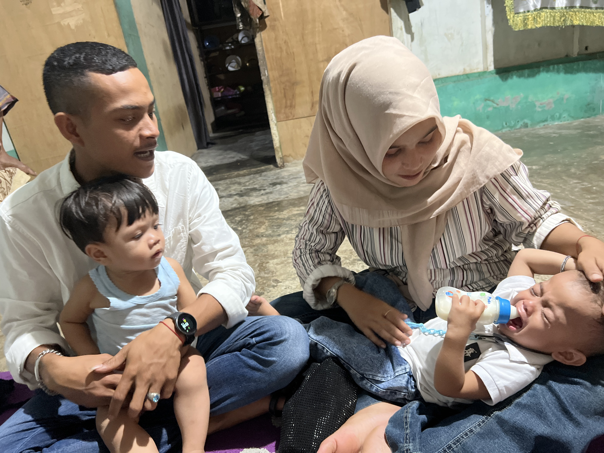 The image depicts a family scene in a simple, indoor setting, likely a home in Lhokseumawe, Indonesia. The environment appears to be a room with concrete flooring and a wall with some peeling paint. In the foreground, a man wearing a white long-sleeved shirt and blue jeans is holding a young child. He is looking at the child with a gentle expression. Beside him, a woman wearing a light brown hijab and a striped shirt is tending to another baby, who is crying and being fed from a baby bottle. The woman’s interaction suggests she is trying to comfort the infant. The children are dressed in casual clothing. The overall atmosphere is one of domesticity and care.