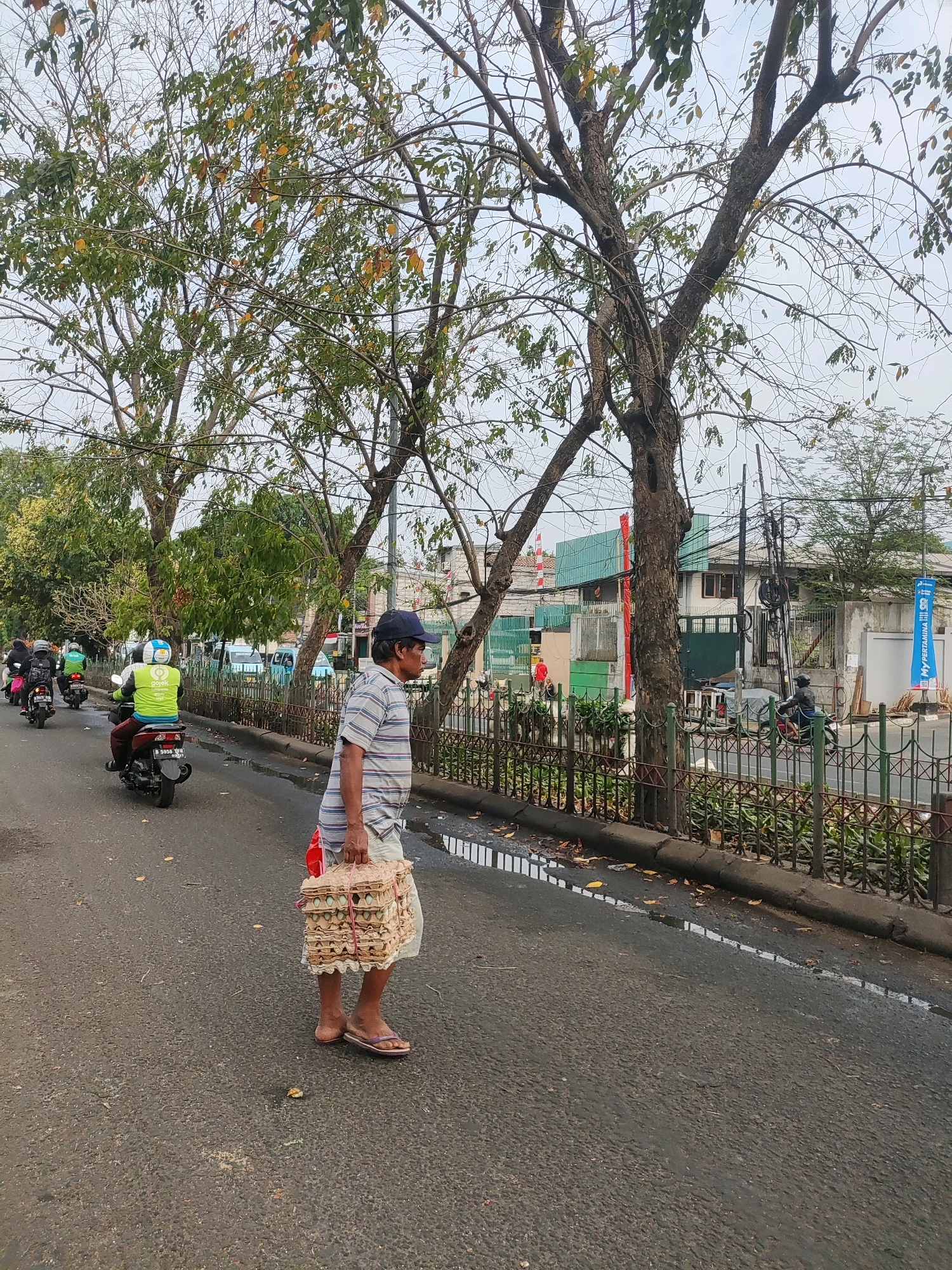 The image depicts a street scene in East Jakarta, Indonesia. In the foreground, a man wearing a blue cap, a striped t-shirt, and light-colored shorts walks on the right side of the road, carrying a large stack of egg cartons in his right arm. He is wearing flip-flops. The road is asphalt and appears wet in places, with puddles reflecting the sky and surrounding environment.
In the background, several motorcycles are seen on the road. One motorcycle in the mid-ground has a rider wearing a bright green vest with the logo "Go\isk" prominently displayed. Another rider behind him also wears a similar green vest. Further down the road, other motorcycles and a light blue vehicle are visible.
The street is lined with trees that have sparse foliage, suggesting it might be a dry season or the trees are naturally bare at this time. A decorative metal fence runs along the right side of the road, separating the street from a sidewalk and buildings. Behind the fence, buildings with green roofs and white walls are visible. There are also utility poles with numerous wires running between them. The sky is overcast.