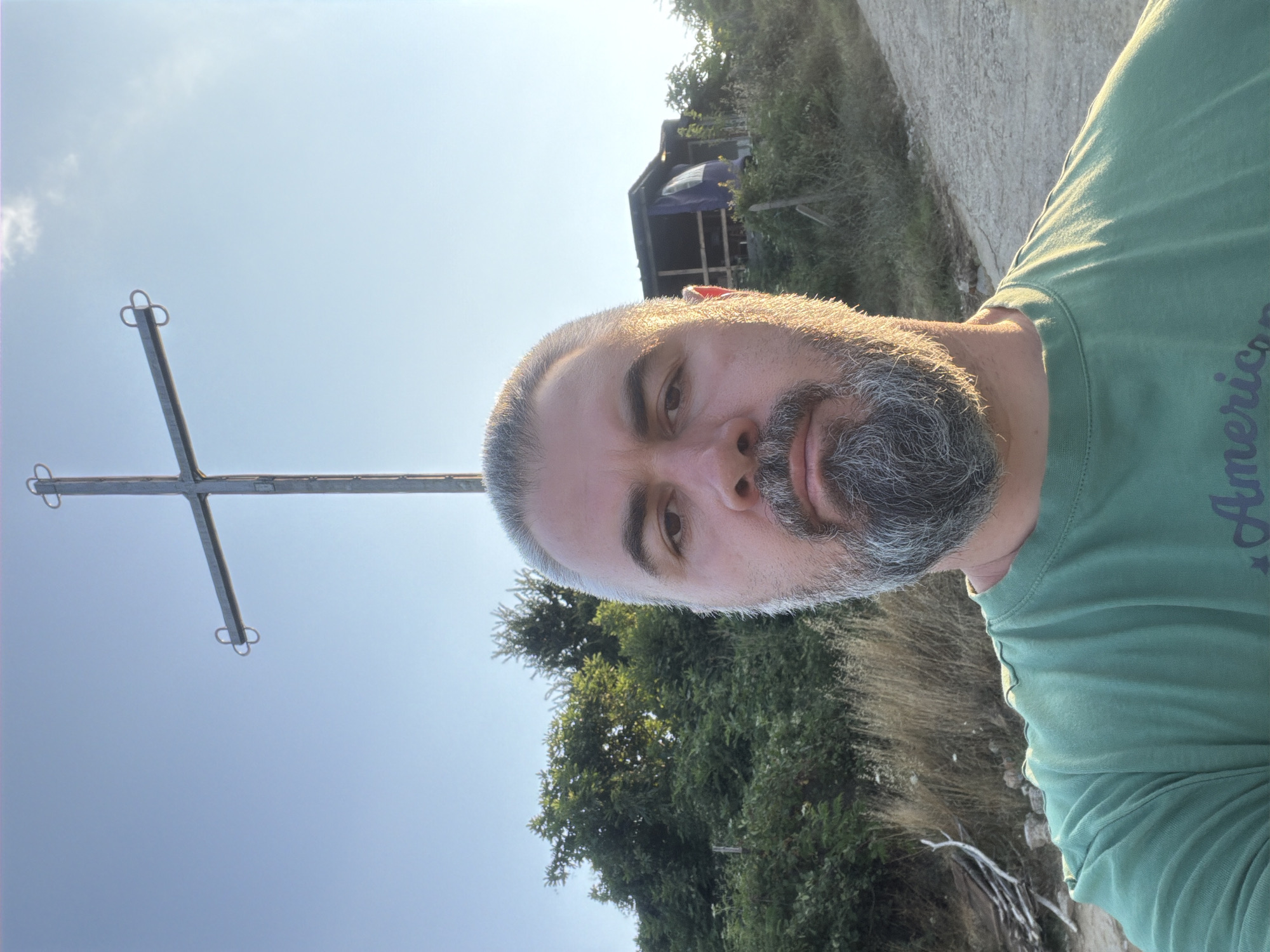A man, with a greying beard and hair, is taking a selfie outdoors. He is wearing a green t-shirt. In the background, a large metal cross stands against a clear blue sky. To the right of the cross, there is a small, rustic structure covered with a blue tarp. The environment appears to be a rural or semi-rural area, with trees and dry grass visible. The lighting suggests it is either early morning or late afternoon. The scene is located in Gniazdovo, Bulgaria.