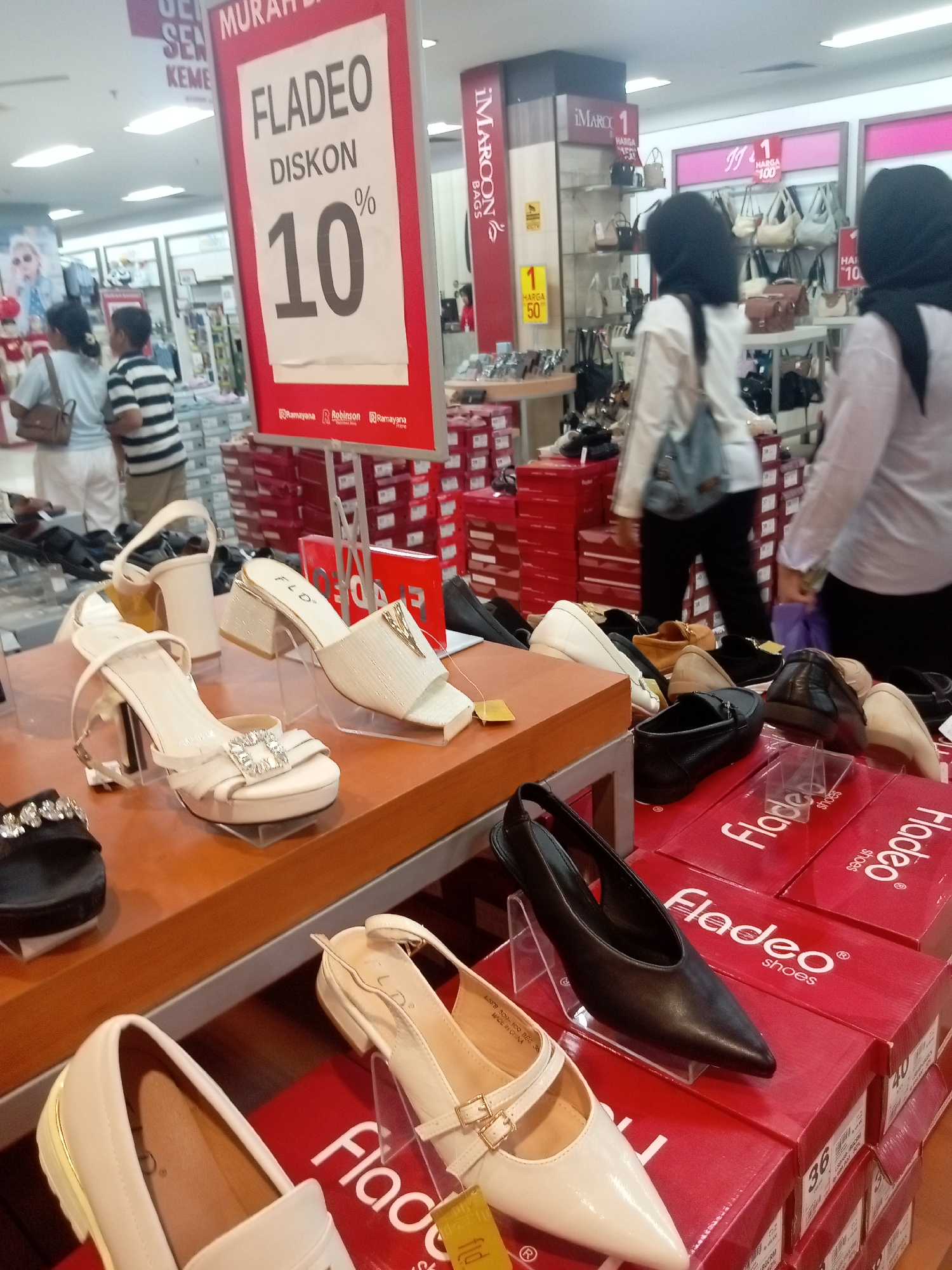The image captures a shoe store display in Batam, Indonesia, likely within a department store like Ramayana, as indicated by signage. In the foreground, several pairs of women's shoes are arranged on a wooden display shelf and on top of red shoeboxes. The shoes include white heeled sandals with decorative buckles, white slingback heels, and a black pointed-toe slingback heel. Behind the shoes, red shoeboxes are stacked high, with "Fladeo shoes" branding visible. In the midground, two women wearing headscarves are browsing the merchandise. One woman, wearing a light-colored top and a blue backpack, is facing away from the camera. Another woman, to the right, is also wearing a headscarf and a light-colored top, her back to the camera. In the background, more shoe displays and racks of handbags can be seen. A large red sign with white text announcing "FLADEO DISKON 10%" is prominently displayed on the left side of the image, indicating a sale. The environment appears to be a well-lit retail space.