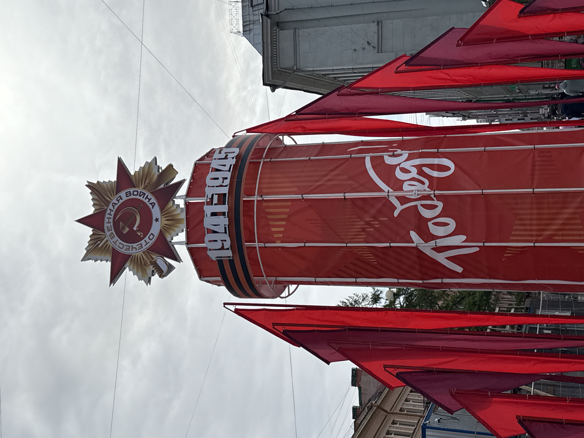 The image shows a street decoration in Saint Petersburg, Russia, related to the commemoration of the Great Patriotic War (World War II). The decoration includes a large red banner with the dates "1941-1945" and a Soviet star with the Order of the Patriotic War emblem. Above and below the banner, there are numerous red flags hung across the street. The banner is also adorned with a white inscription. Buildings can be seen along the sides of the street, indicating an urban setting. The sky above is overcast.