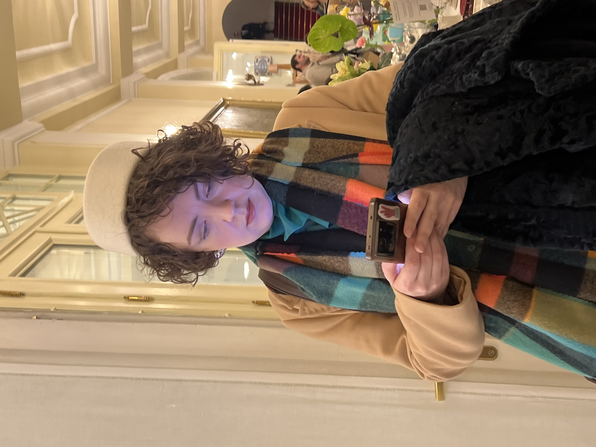 A woman is shown in an indoor setting. She wears a white hat, a tan jacket, and a multi-colored scarf. She is using a smartphone. The setting appears to be an ornate interior with decorative ceilings and large windows. There are other people in the background, as well as flowers and table settings suggesting a gathering or event. The location appears to be in Saint Petersburg, Russia, due to the architectural style reminiscent of buildings in the city, however, this location cannot be confirmed without more information.