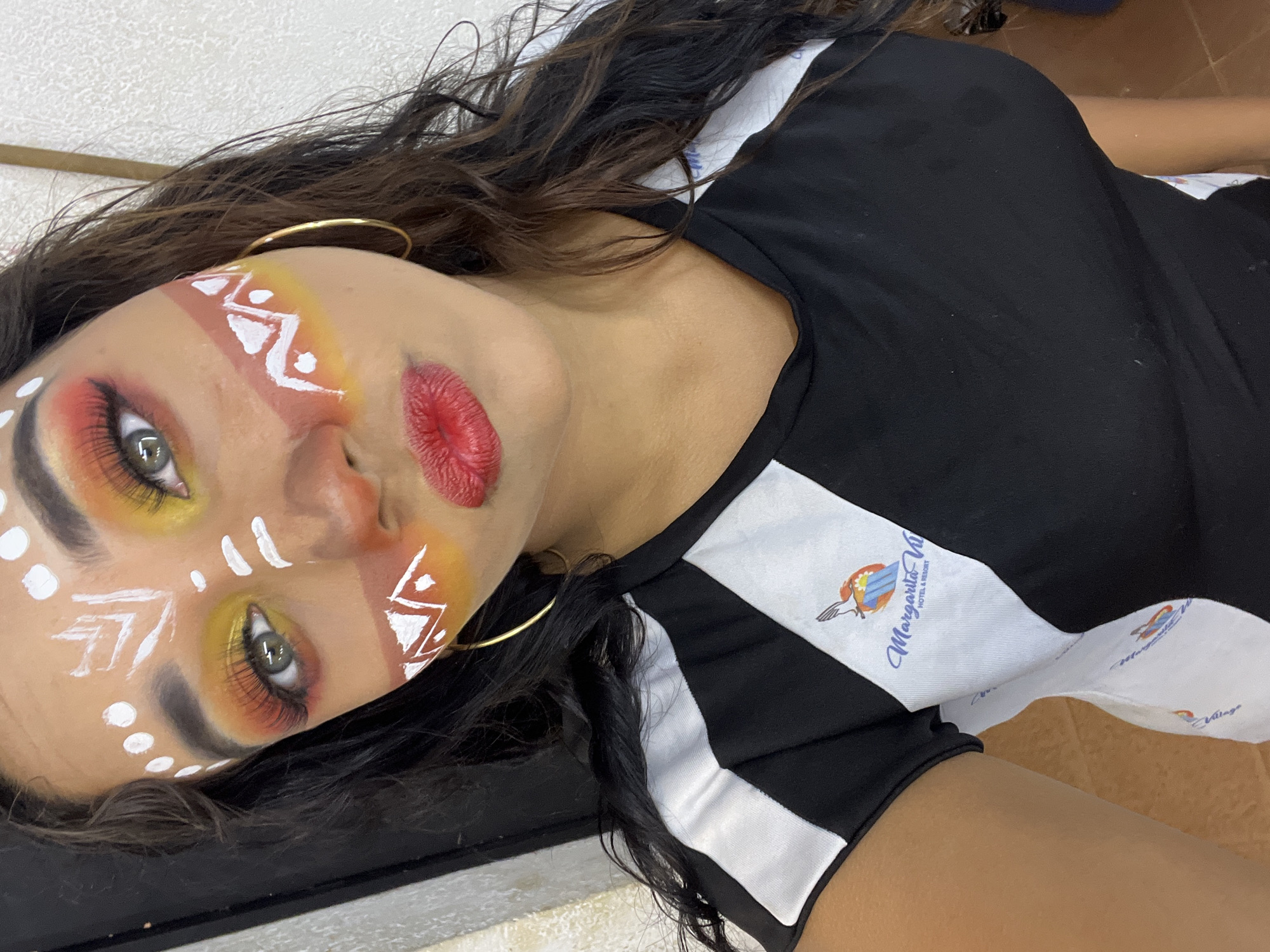A woman with painted face and red lips lies at an angle, wearing gold hoop earrings and a black top with white stripes, at the Margarita Village Hotel & Resort in Porlamar, Venezuela. The face paint includes white geometric patterns and yellow-orange shading near the eyes. The top has the Margarita Village Hotel & Resort logo on the white stripes.