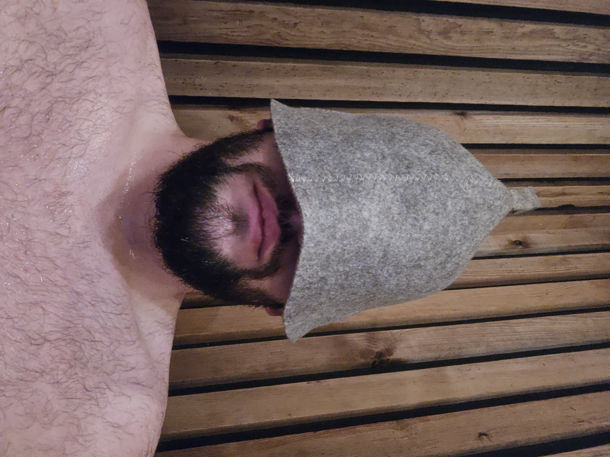 A close-up, high-angle shot captures a person, likely male due to visible facial and body hair, relaxing in a sauna in Sofia, Bulgaria. The individual is seen from the chest up, lying on a wooden slatted surface, presumably a sauna bench. Their skin, particularly on the shoulder and chest area, is flushed and glistening with sweat or moisture, indicating a hot environment.
The person's face is partially obscured by a light grey, conical felt sauna hat, which covers the upper part of their head and eyes. A dark beard and mustache are prominent around the mouth and chin, with the lips appearing slightly parted. The hat features visible white stitching along its edge, adding a textural detail.
The background consists entirely of horizontal wooden planks, typical of sauna construction, suggesting a warm and rustic setting. The wood appears to be a natural medium-brown tone with subtle variations. No specific time of day or weather is discernible from the indoor setting, but the visible sweat and sauna hat strongly imply an activity of thermal relaxation. No text is visible in the image. The overall impression is one of peaceful respite in a heated environment.